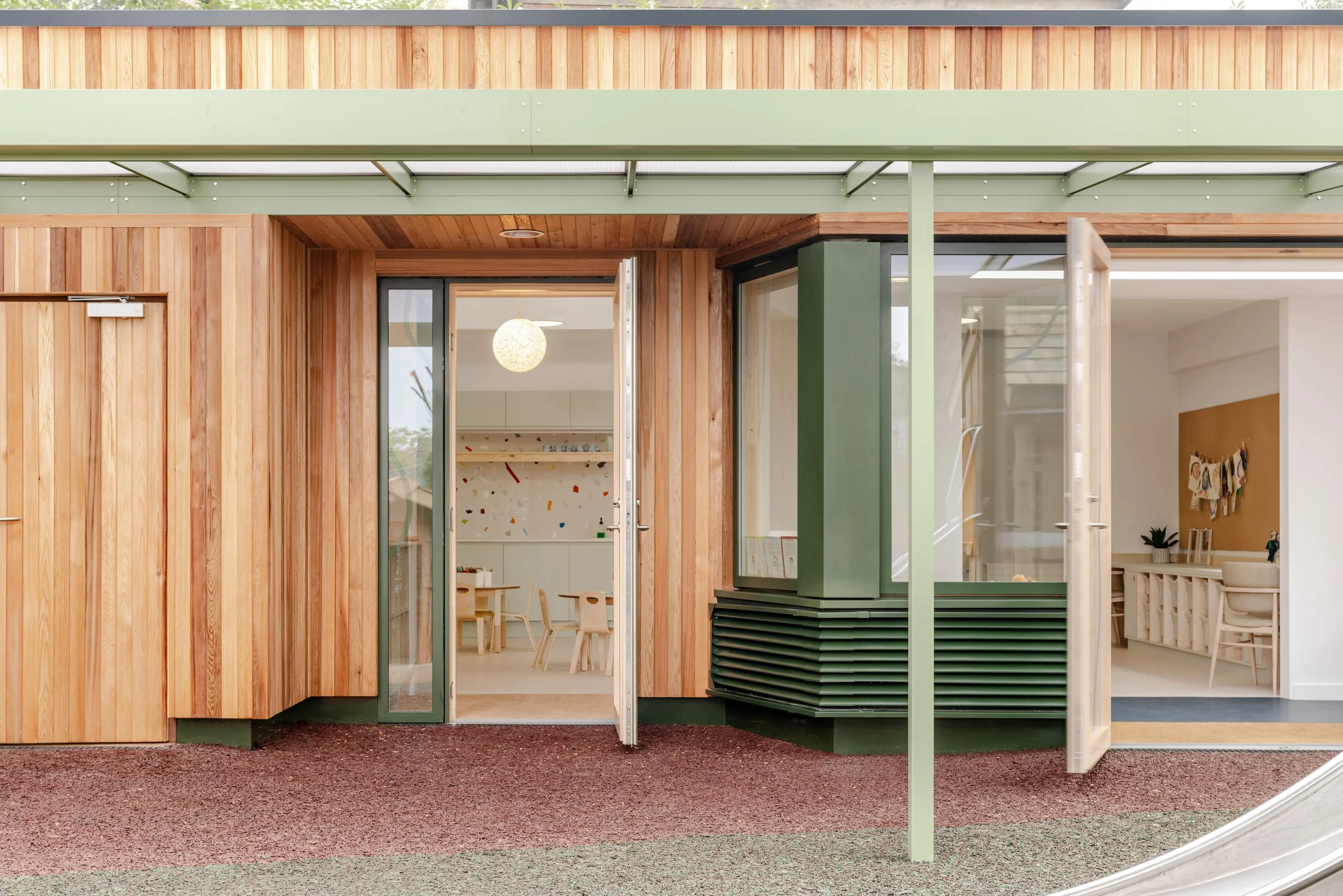 Timber-clad early years school building with open classroom doors connecting indoor learning spaces to a covered outdoor play area designed for pupil wellbeing and flexible learning.