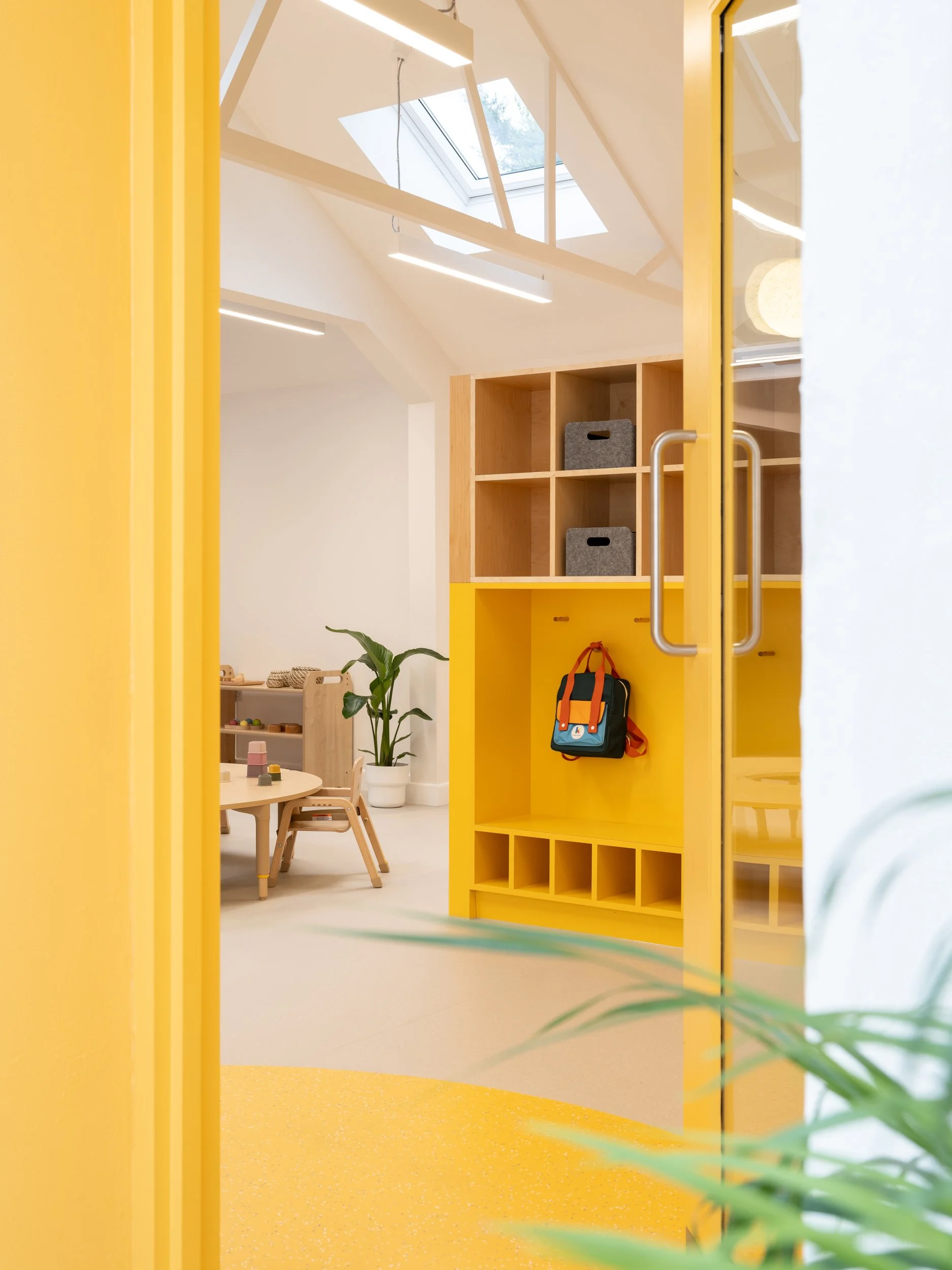 Early years classroom entrance with bright yellow joinery, child-height storage, and naturally lit learning spaces designed to support independence and welcoming educational environments.