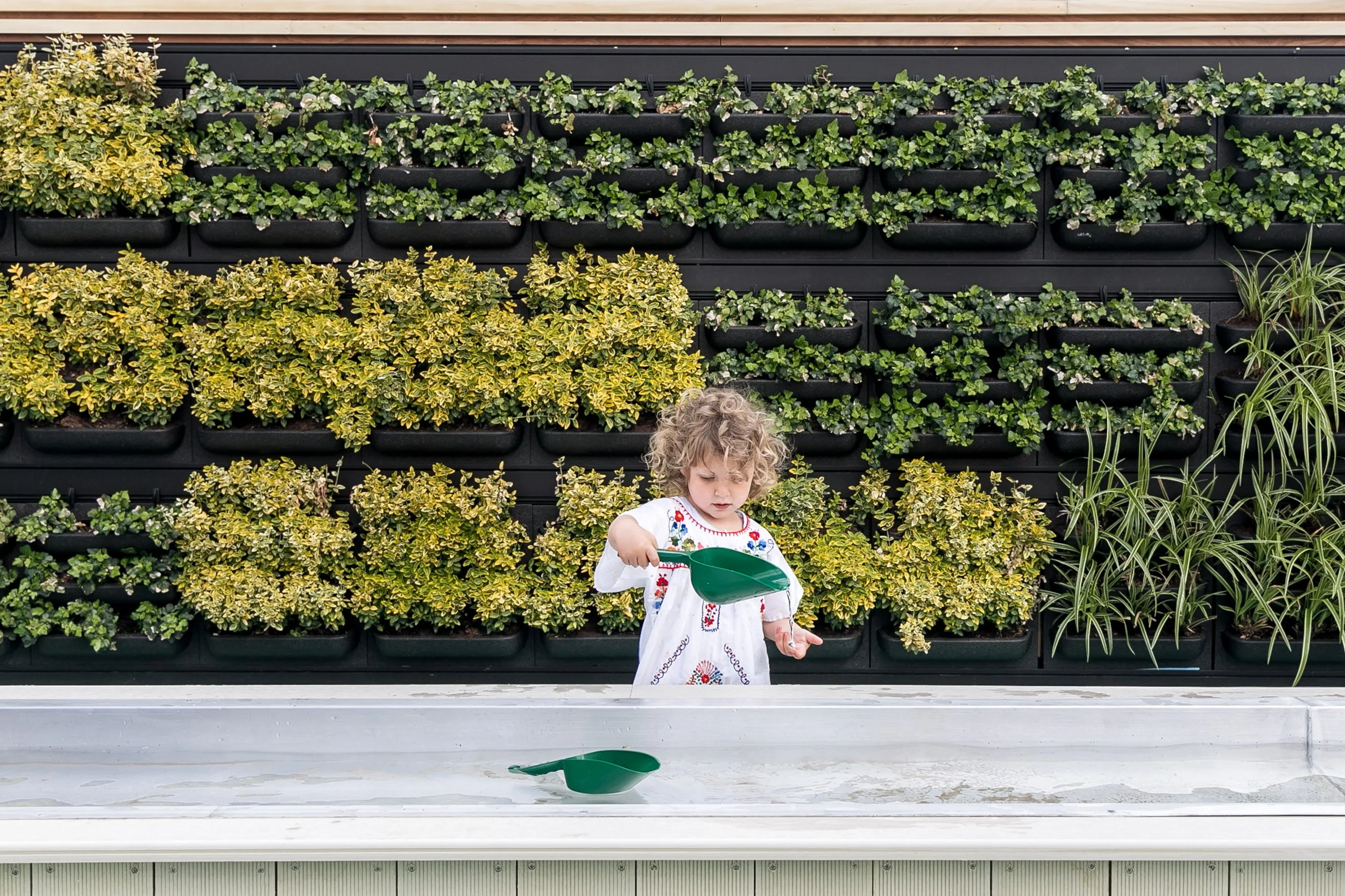 Child engaging with water play in an outdoor learning space featuring a living green wall, integrated planting, and nature-based play elements designed for early years education.