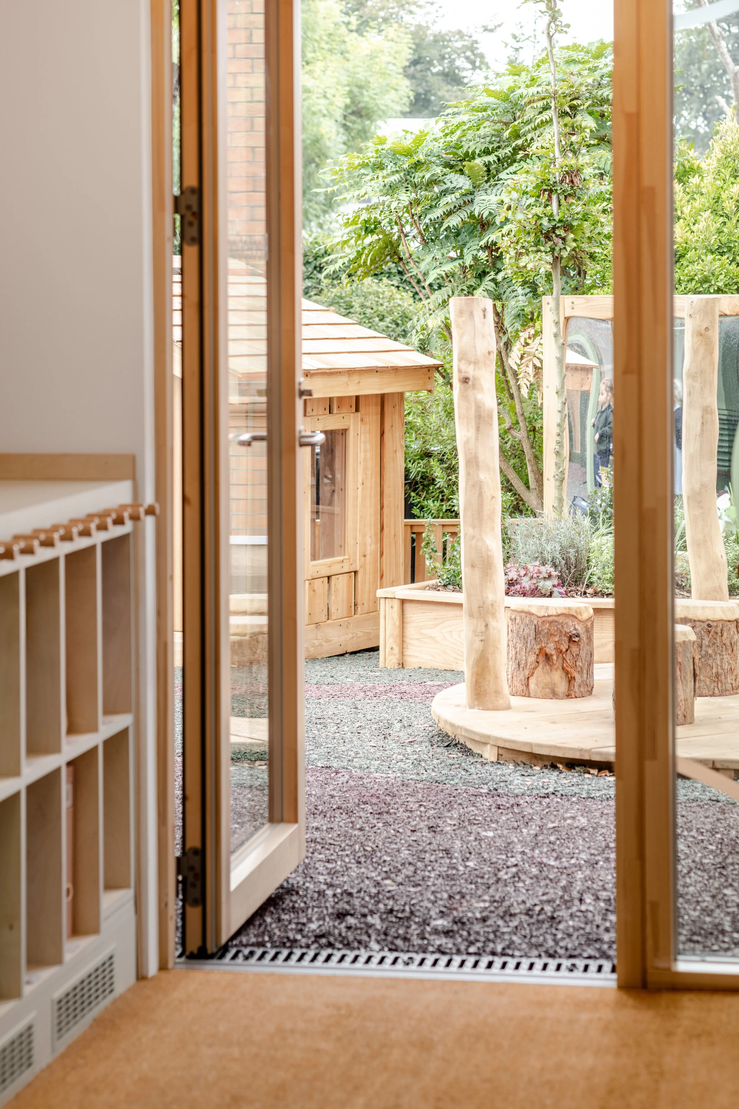 School classroom interior with open timber-framed doors leading to a natural play courtyard featuring timber planting beds, outdoor learning areas, and child-centred landscape design.