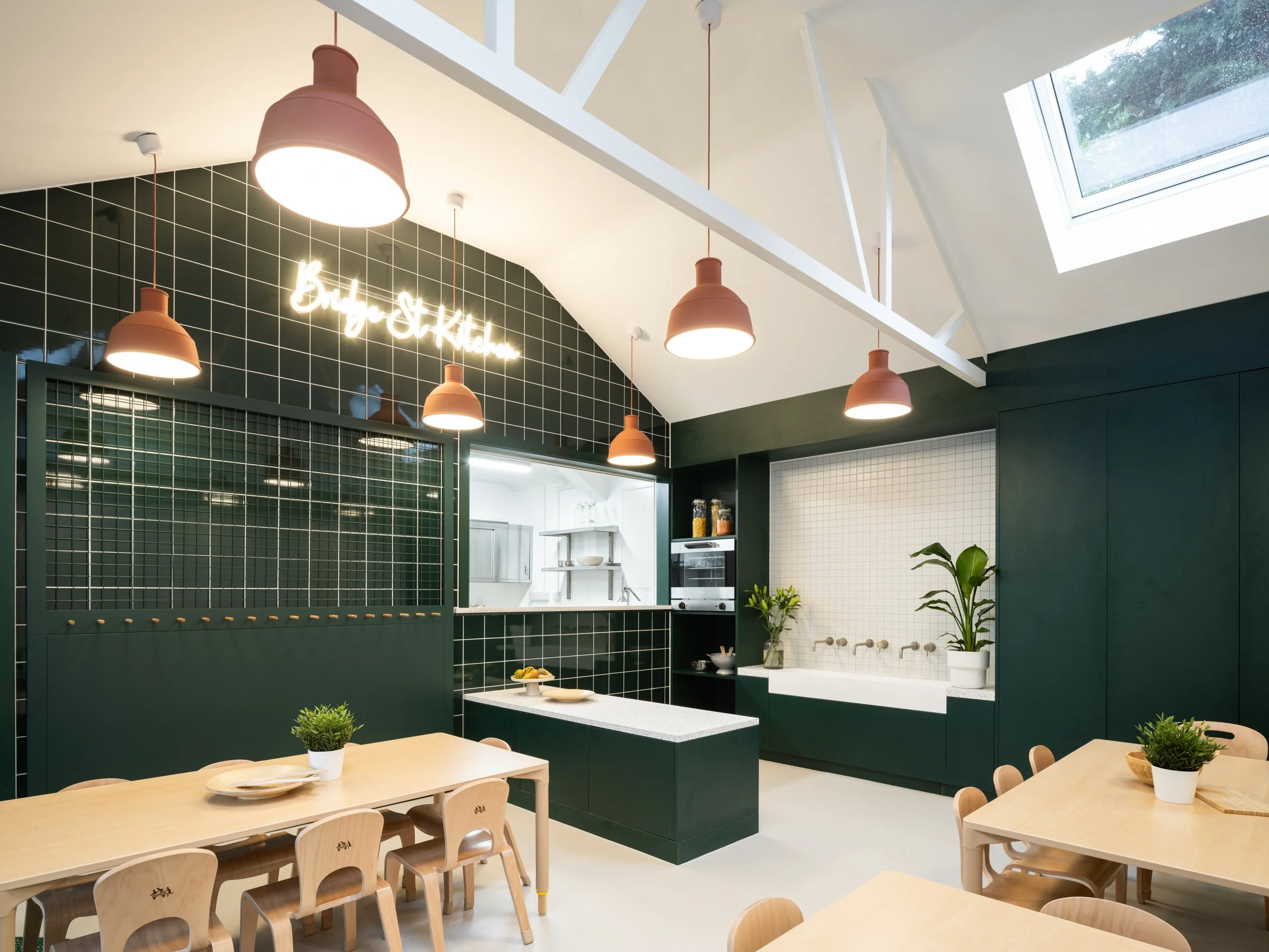 Early years kitchen and dining space with child-sized tables, dark green tiled joinery, and rooflights designed to support shared meals, practical learning, and community-focused education.