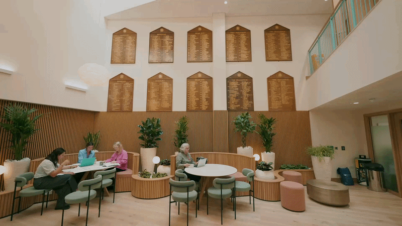 Inside a modern lobby with high ceilings, wood paneling, and large hanging boards on the wall, four women are sitting at tables using laptops, surrounded by potted plants and comfortable seating.