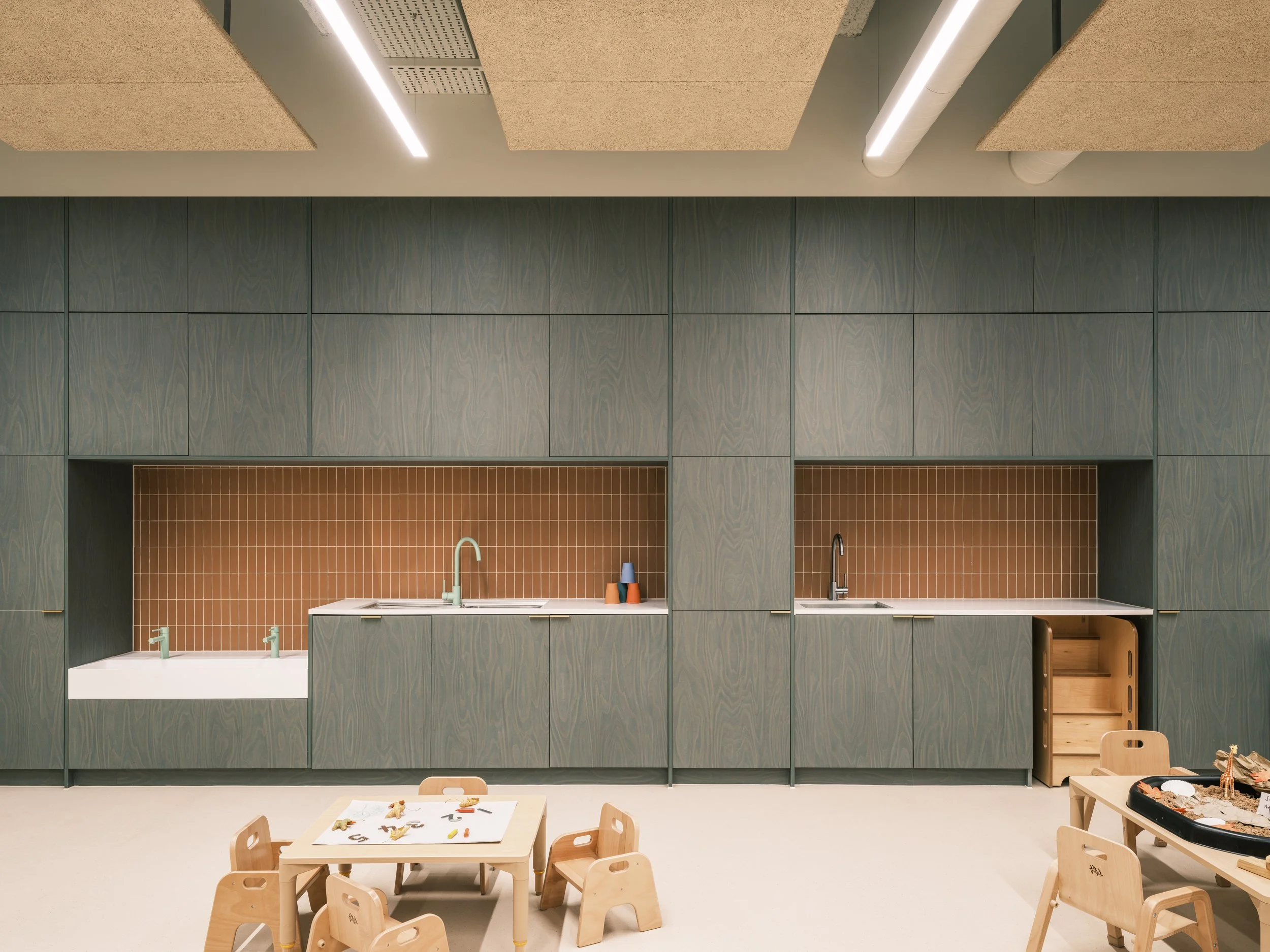 Contemporary early years learning environment featuring bespoke timber cabinetry, integrated child-height sinks, warm terracotta tile splashbacks, acoustic ceiling panels, and Montessori-inspired furniture designed for independent learning and play.
