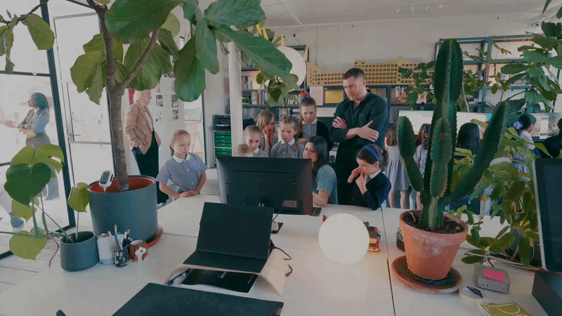 A group of children and a teacher gathered around a computer in a classroom filled with large plants.