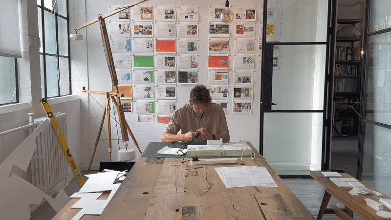 Animated scene of an architect working at a large studio desk surrounded by design sketches, presentation boards, architectural models, and creative workspace materials in a contemporary office setting.