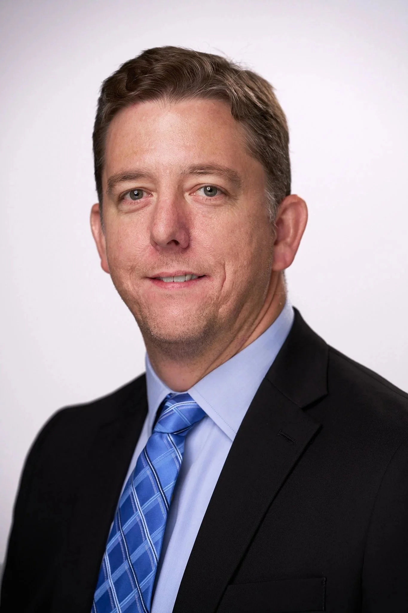 Close-up headshot of a smiling man in a business suit with a blue tie, against a plain white background.