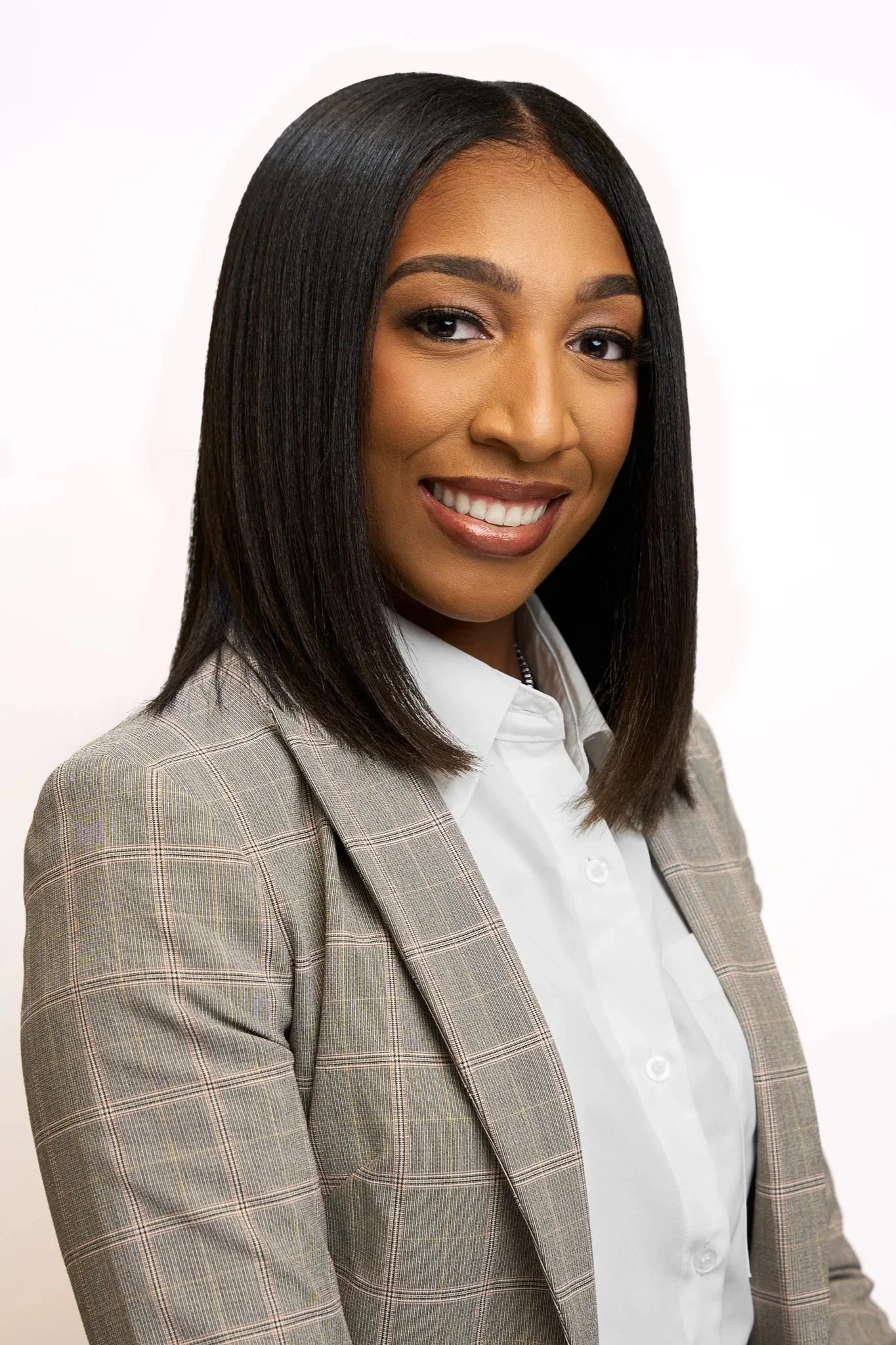A professional woman with shoulder-length black hair, wearing a beige plaid blazer and white shirt, smiling against a plain white background.