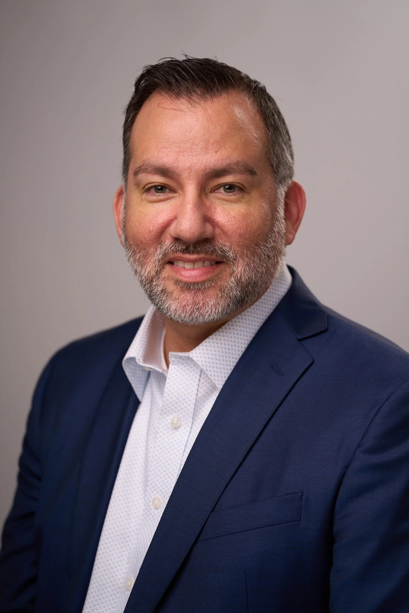 Portrait of a middle-aged man with dark hair and a beard, wearing a navy blazer and a white patterned shirt, smiling slightly against a plain gray background.
