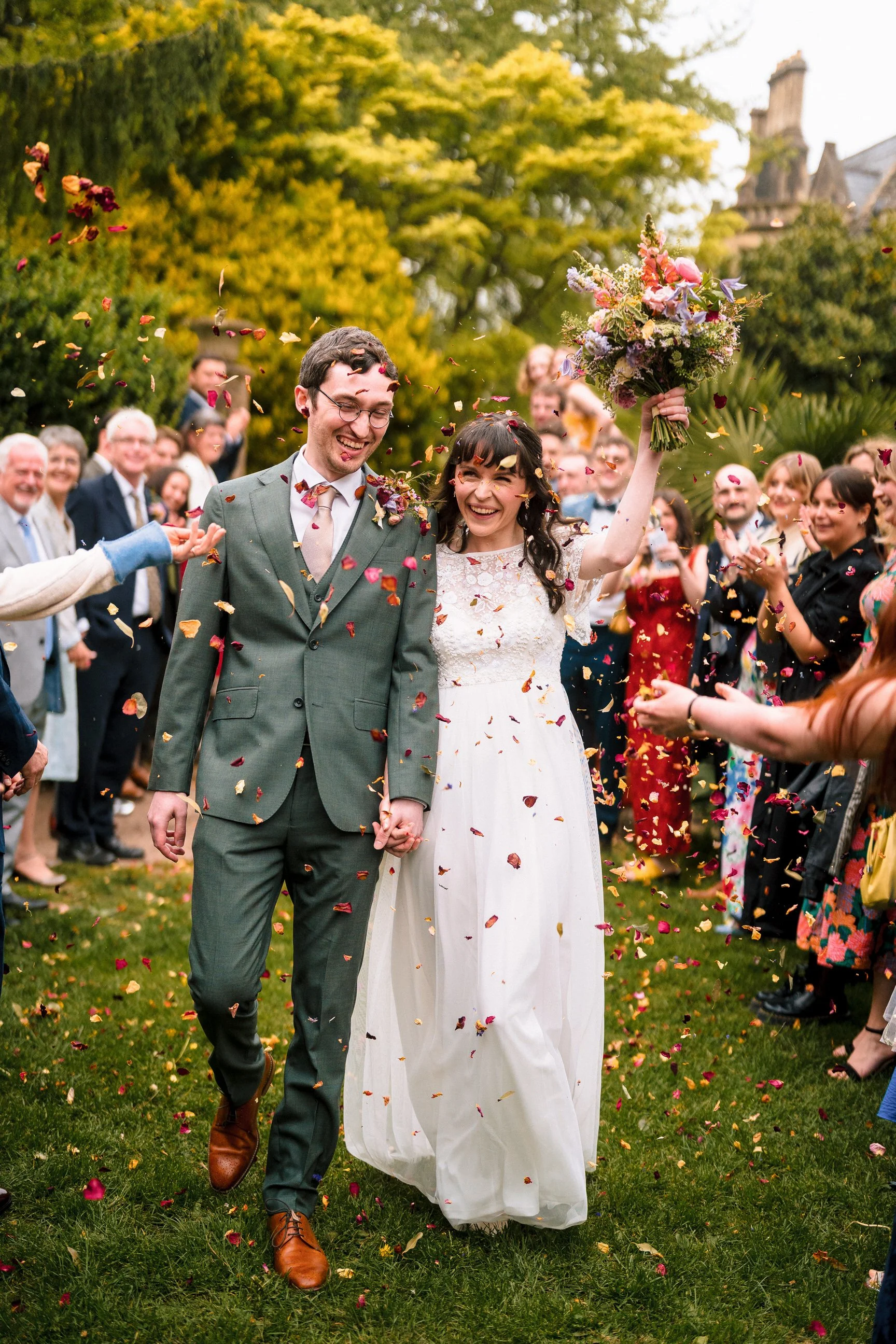 Newlywed couple walking through a garden as guests toss colorful flower petals, with the bride holding a bouquet.