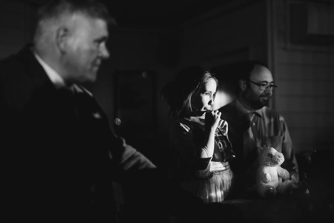 Black-and-white photo of a young girl holding a teddy bear between two adults in a dimly lit room