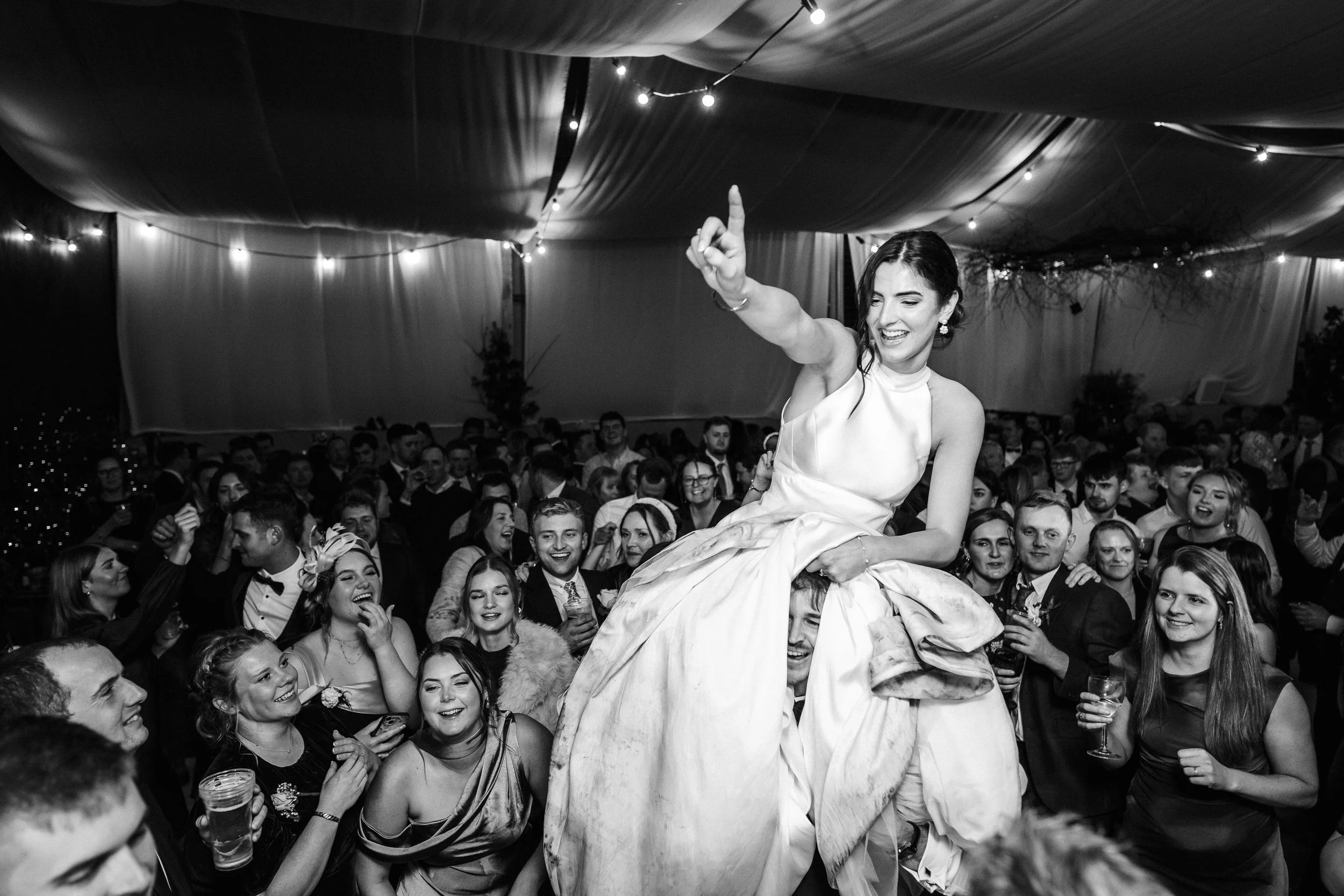 Bride in a white wedding dress raised above a cheering crowd at a reception under string lights