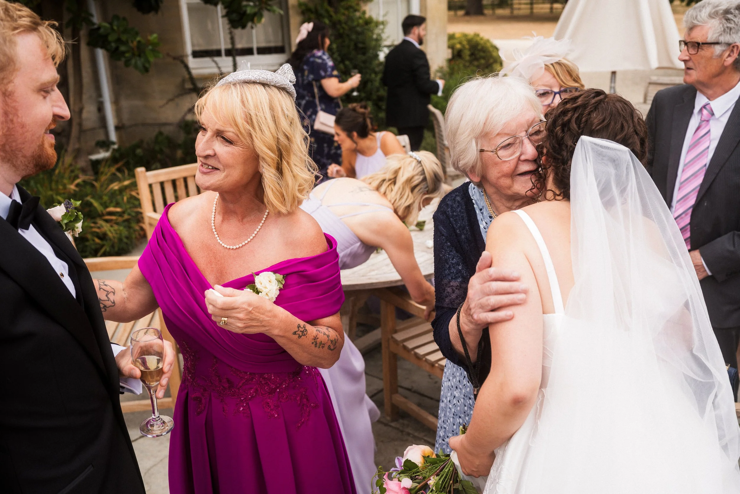 Bride in a white wedding dress and veil embraces an older guest while wedding guests mingle outdoors.