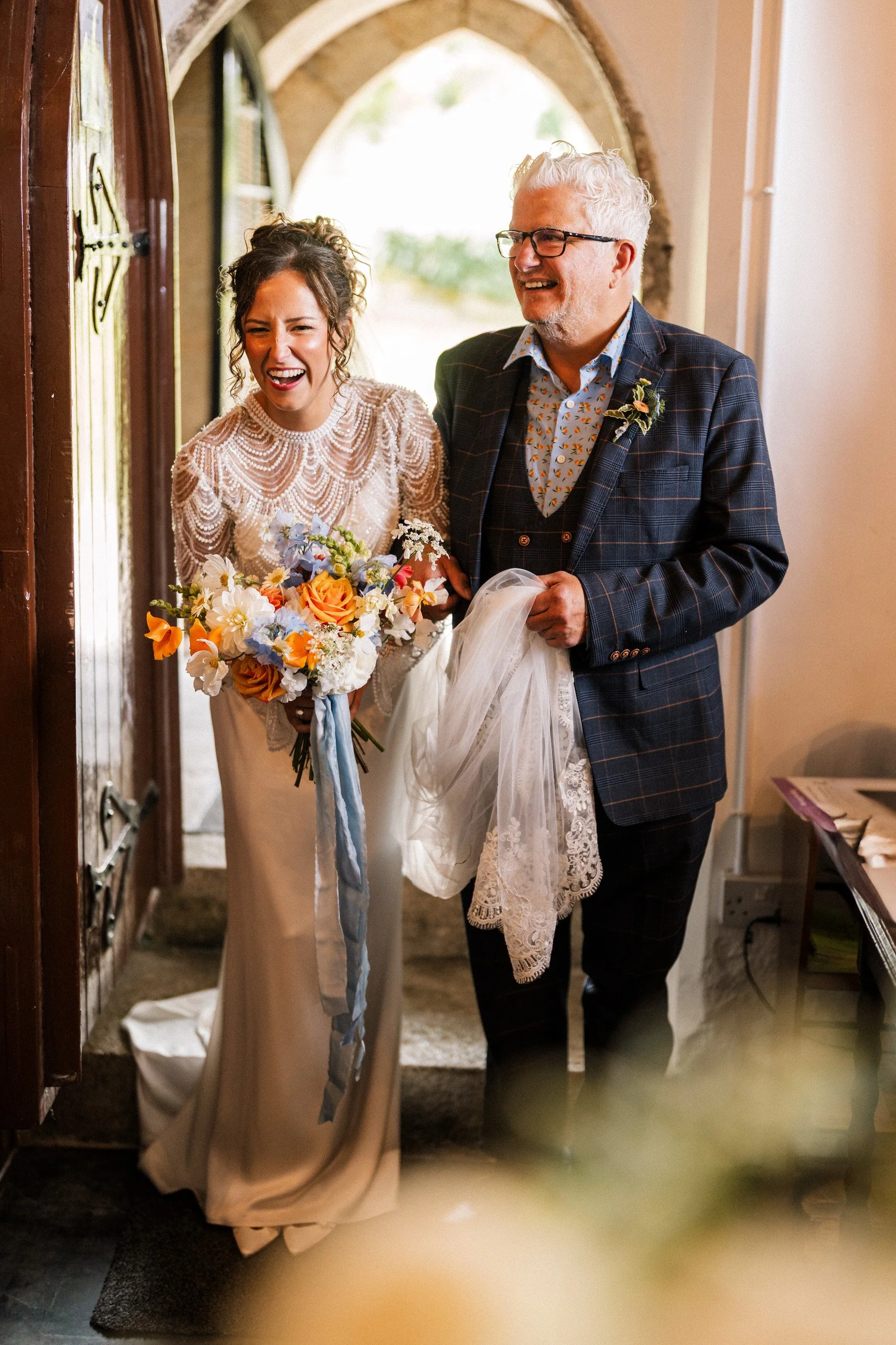 Smiling newlywed couple standing in a doorway, with the bride holding a colorful flower bouquet and the groom wearing a plaid suit.