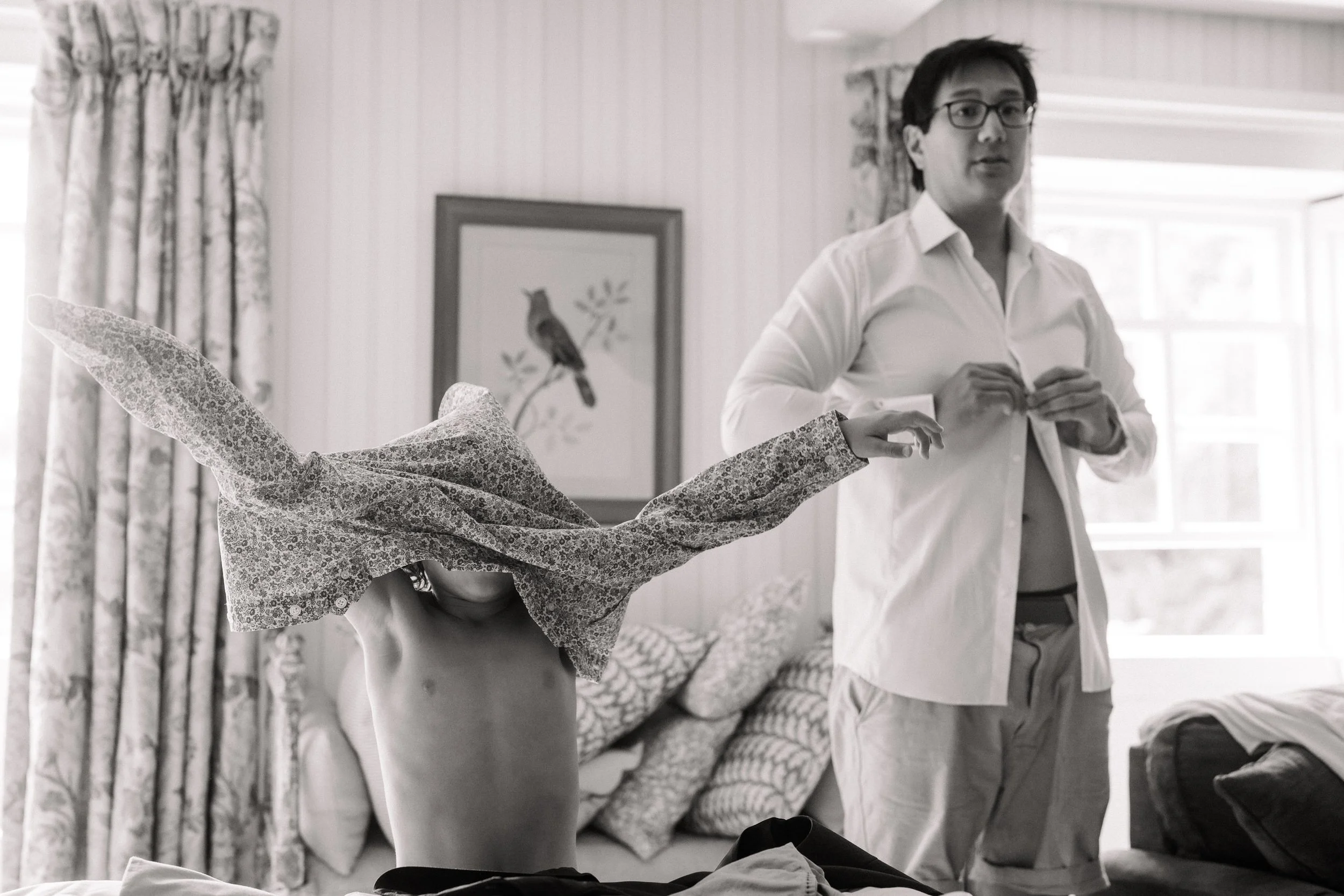 Black-and-white photo of a man in glasses and a white shirt helping another person remove a patterned shirt in a bedroom.
