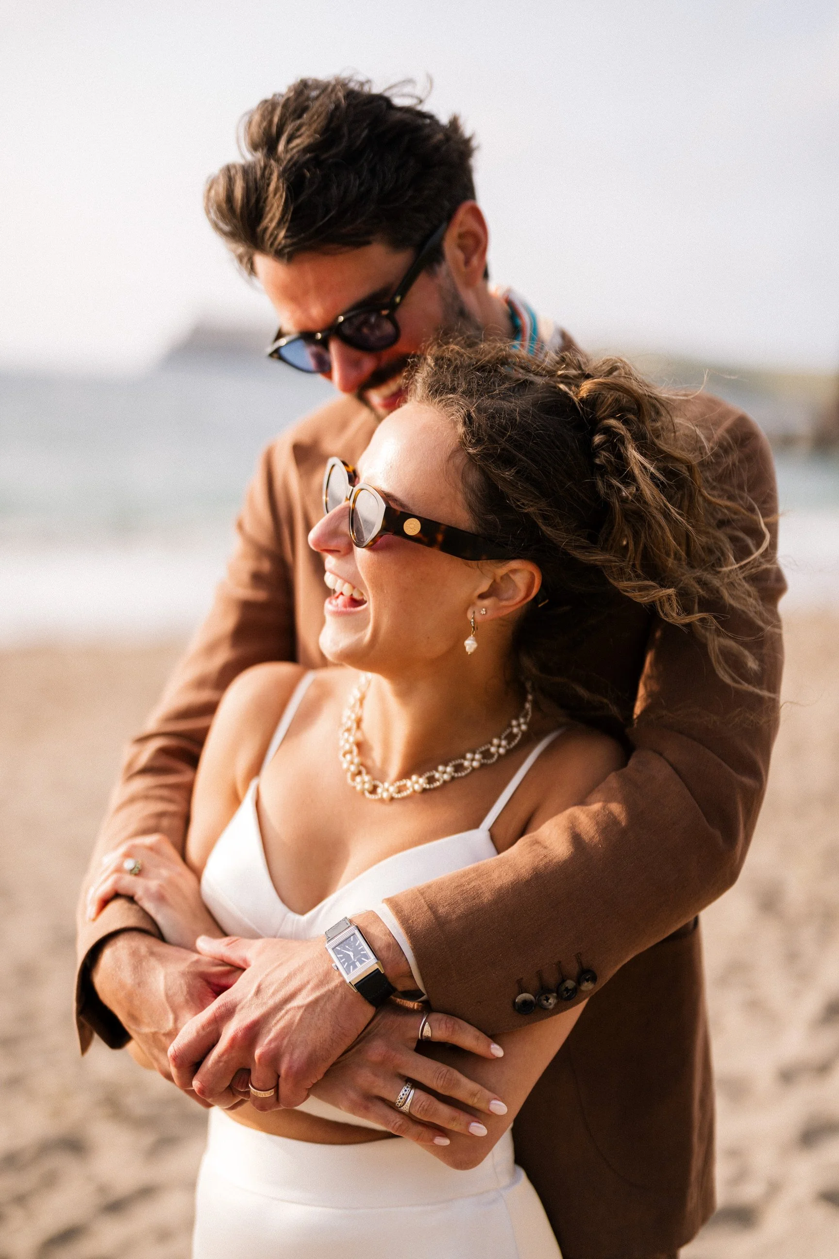 Smiling couple embracing outdoors, with the woman wearing sunglasses, pearl jewelry, and a white dress.