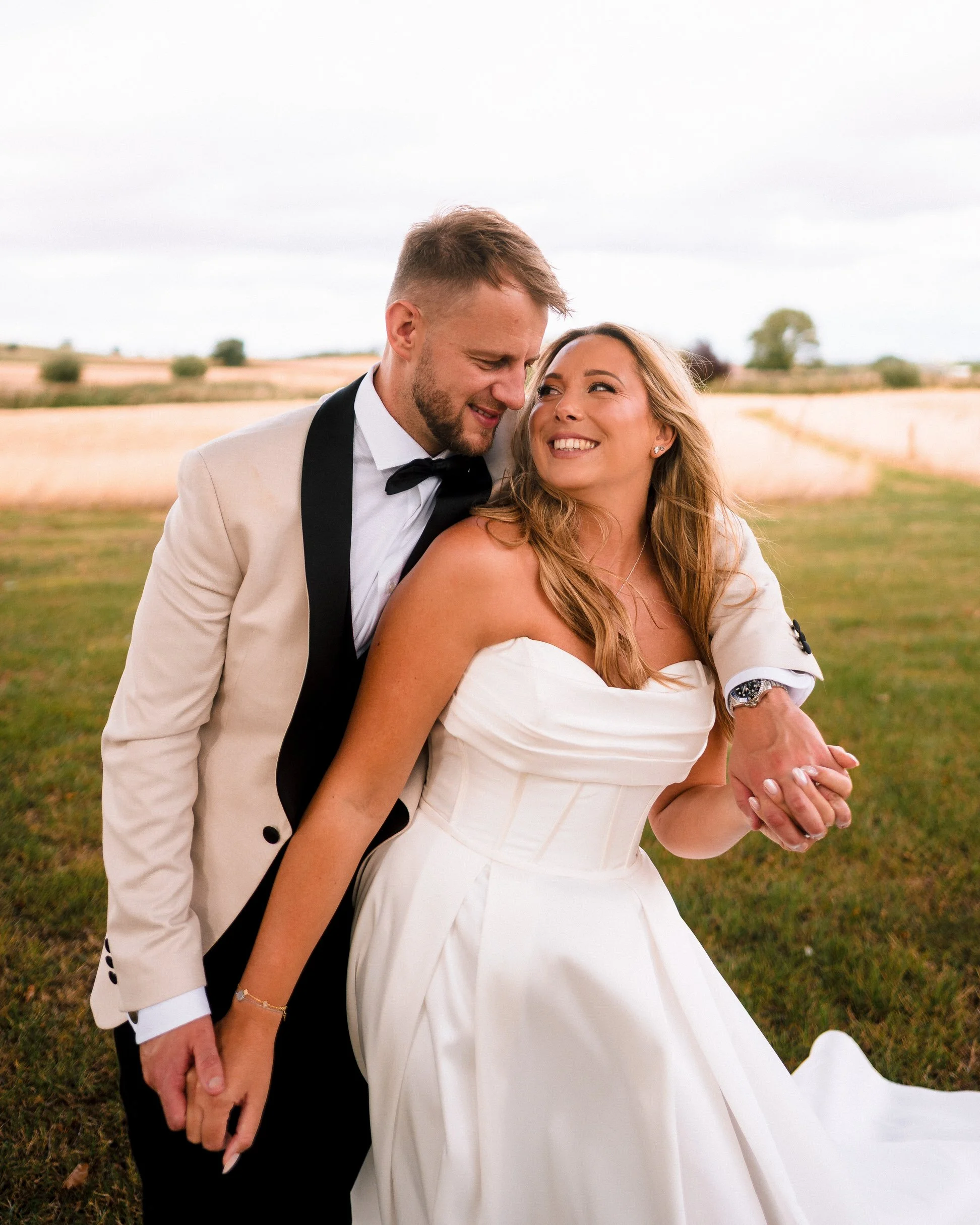 Smiling newlywed couple embracing in a sunlit field, wearing a white wedding suit and white dress.