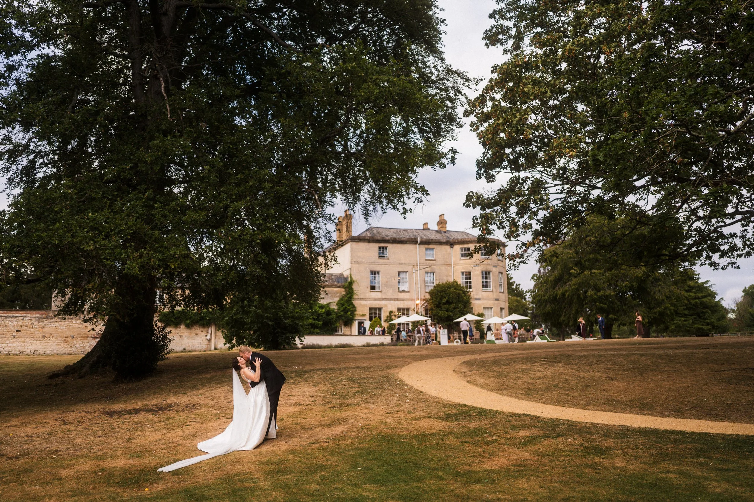 Bride and groom embracing on the lawn of a historic stone wedding venue, with guests and umbrellas in the background.