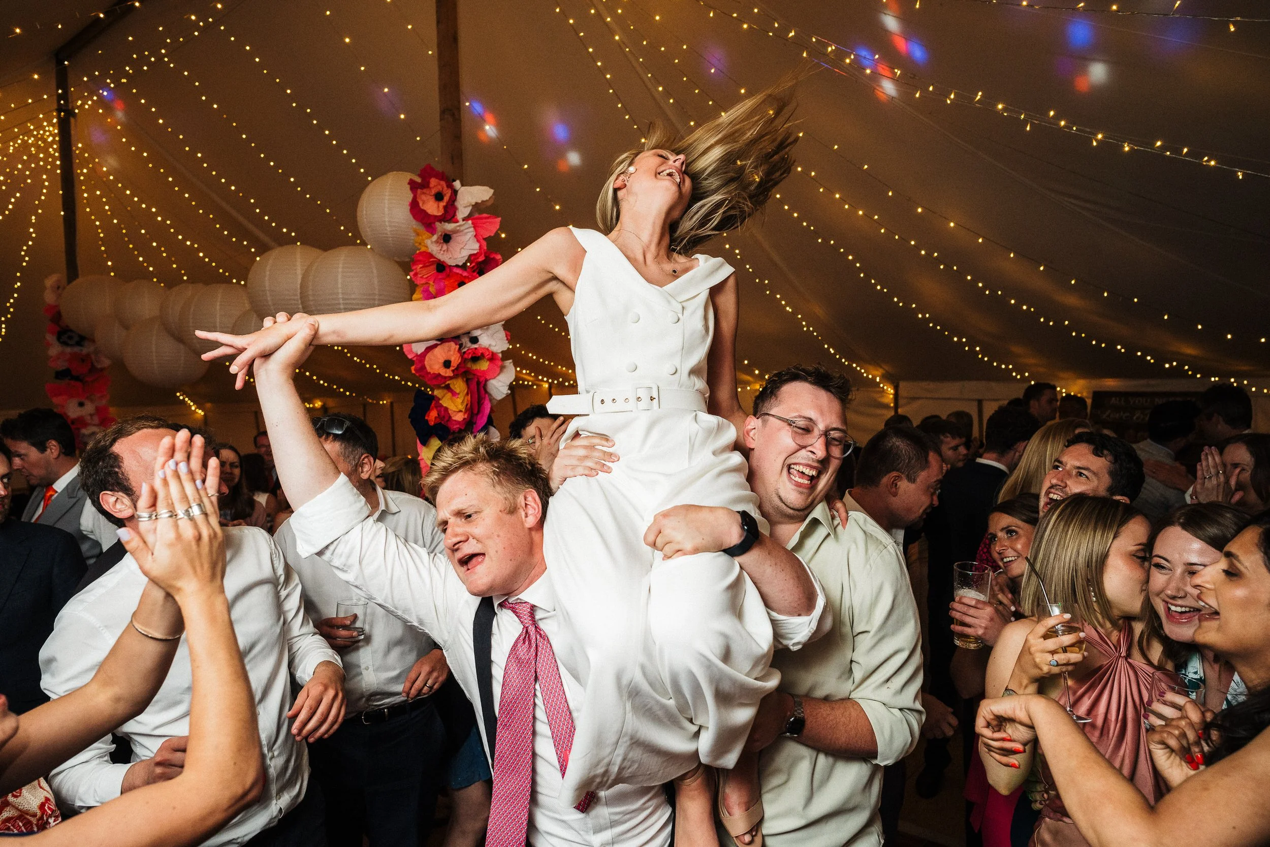 Joyful wedding guests lift a laughing woman in a white dress beneath twinkling string lights.