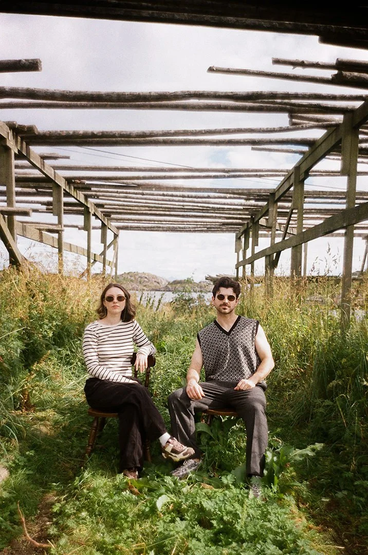 Two people wearing sunglasses sit on chairs beneath a weathered outdoor trellis surrounded by tall grass and wild vegetation.