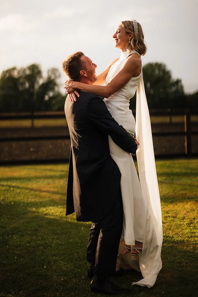 Bride in a white wedding dress embraces her groom in a black suit outdoors at sunset.