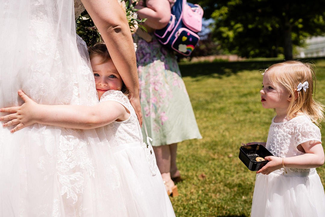 Two young flower girls in white dresses at an outdoor wedding, one hugging the bride and another holding a black ring box.