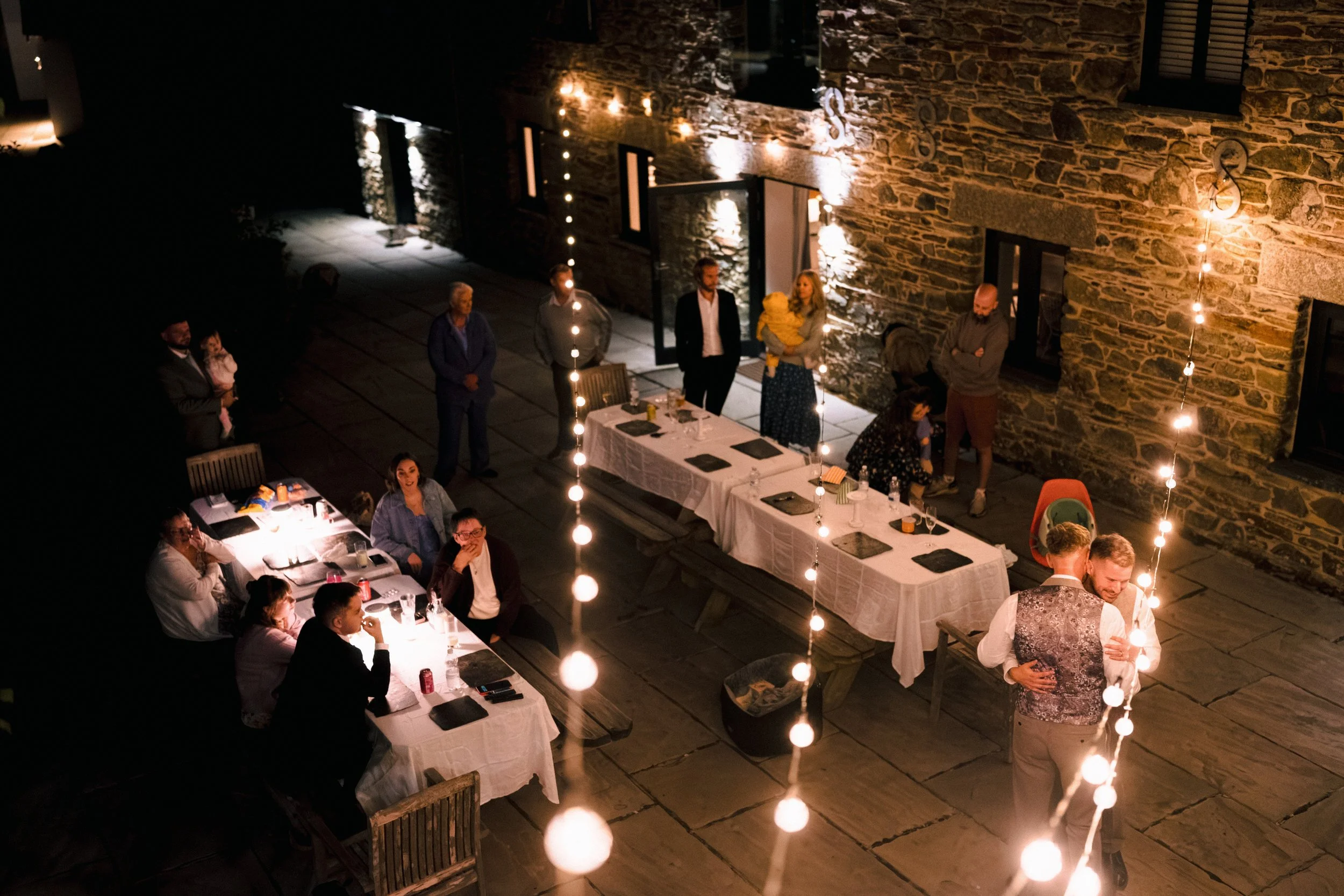 Guests dining and dancing at a warmly lit outdoor wedding reception beside a rustic stone building.