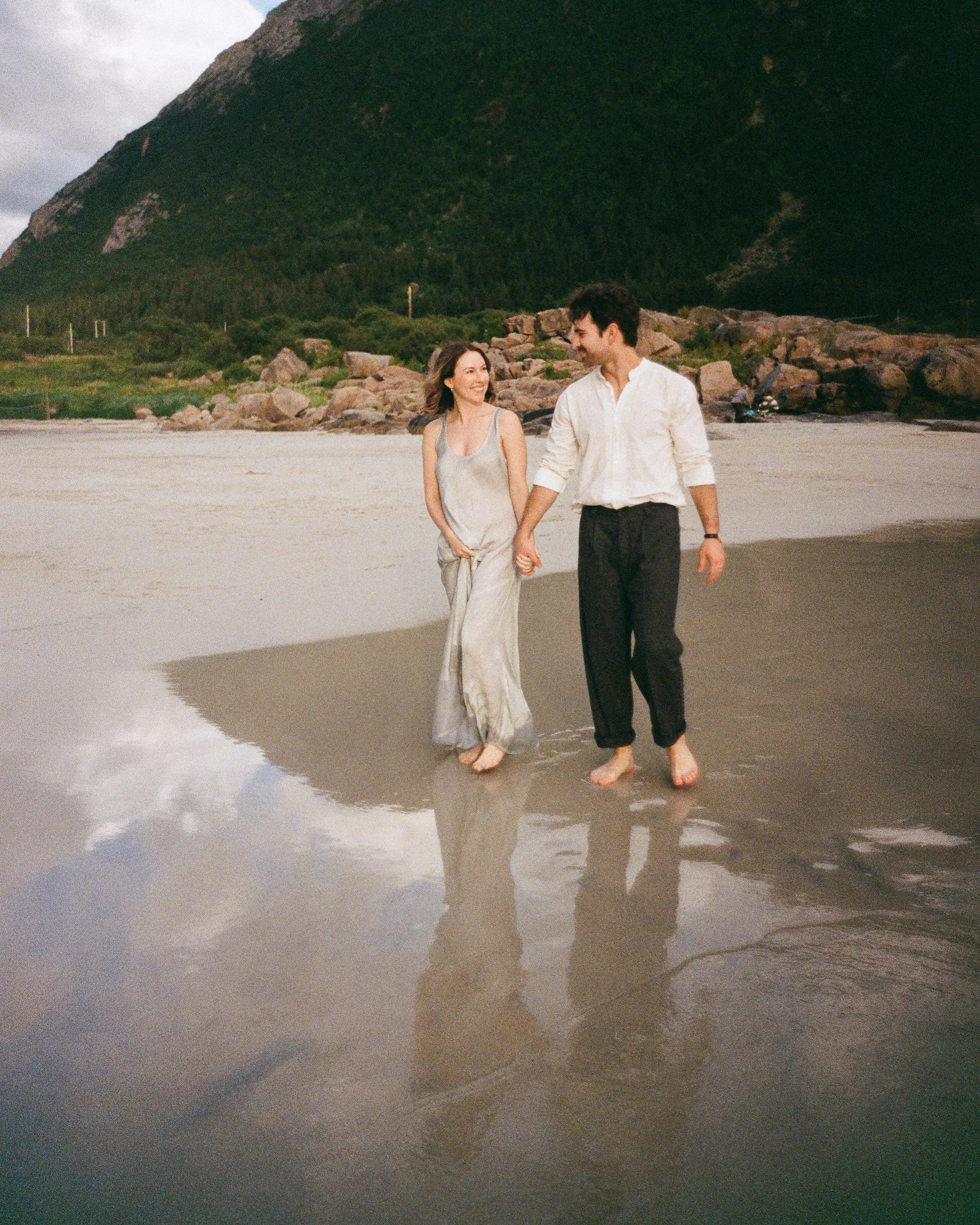 Couple holding hands while walking barefoot in shallow beach water beneath a forested mountain