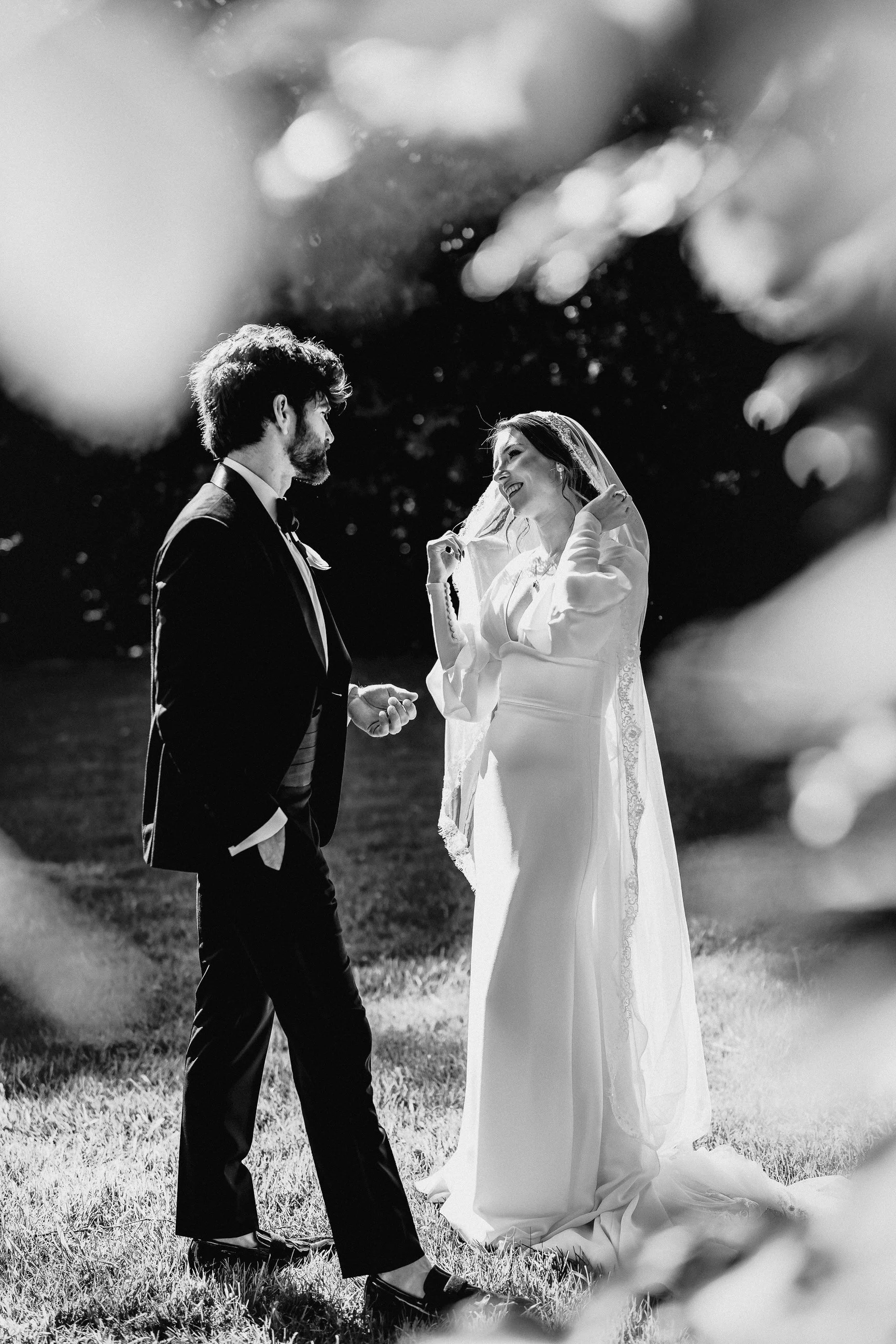 Black-and-white outdoor wedding portrait of a groom holding hands with a smiling bride in a veil and long-sleeved gown.
