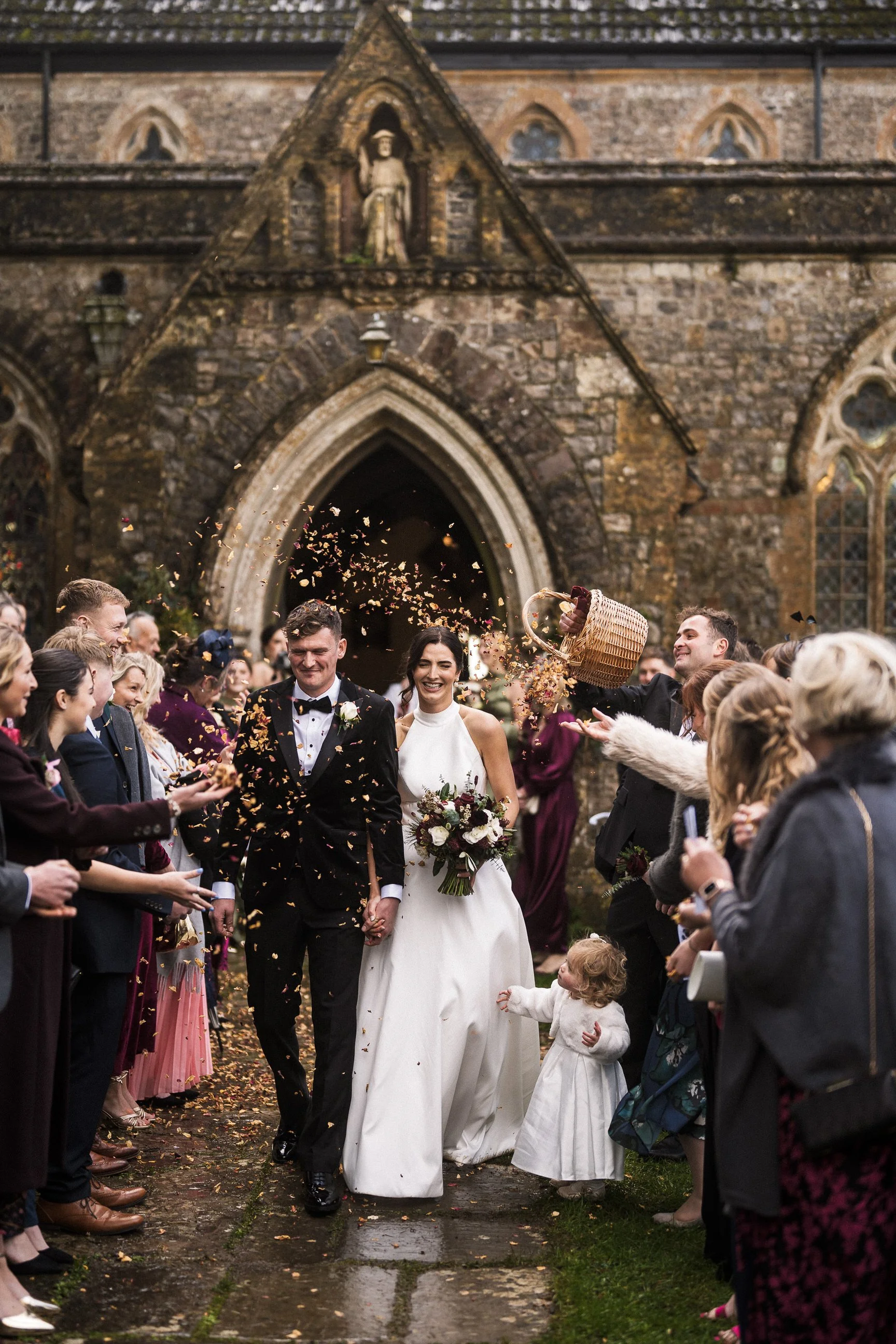 Newlywed bride and groom walk through cheering guests outside a historic stone church as flower petals are thrown.