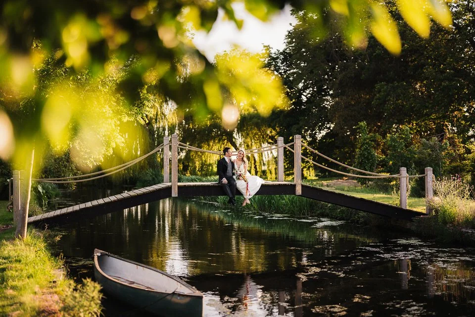 Bride and groom sitting on a wooden footbridge over a scenic garden pond with a canoe in the foreground