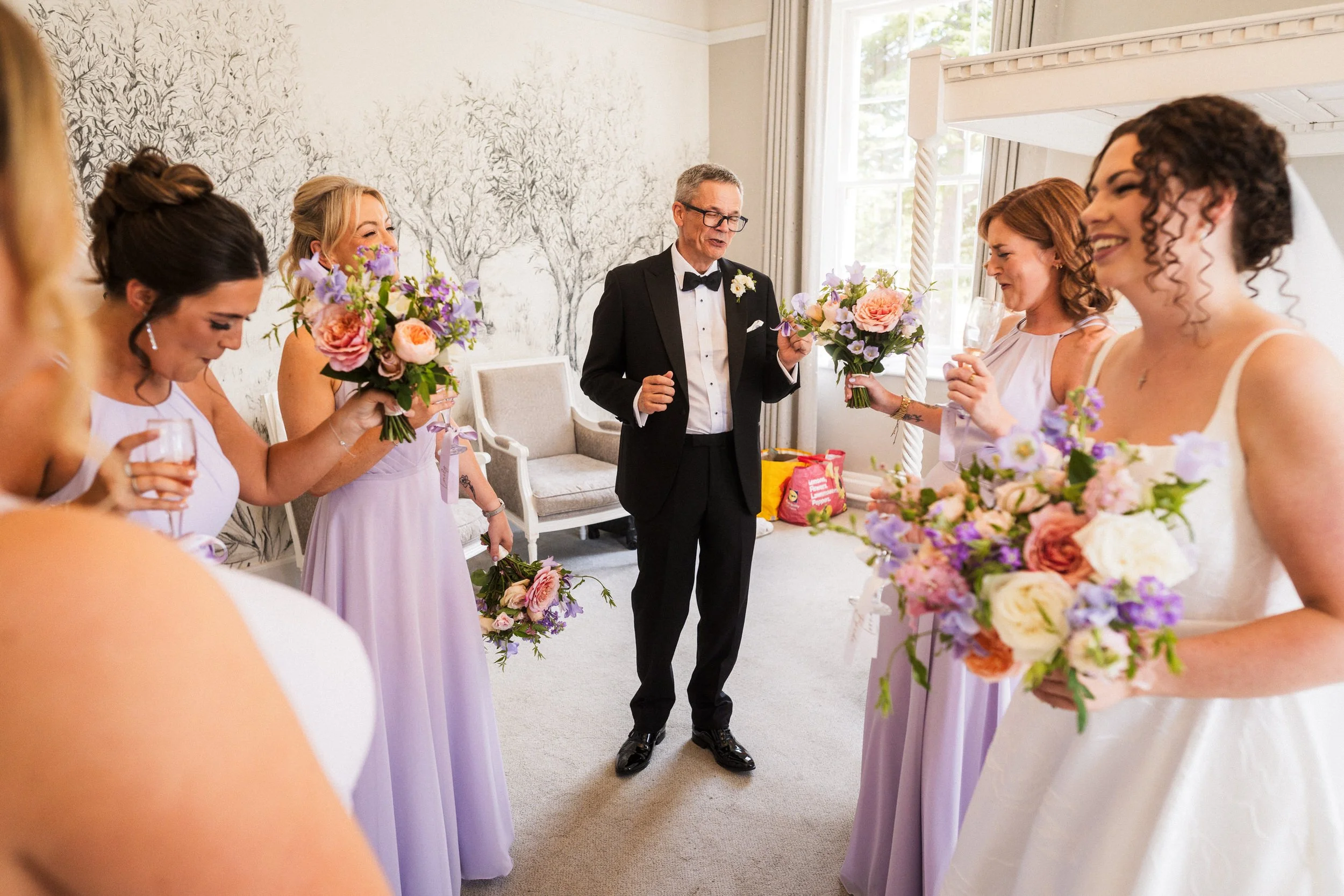 Bride, bridesmaids, and a man in a tuxedo celebrate indoors with colorful flower bouquets and champagne.