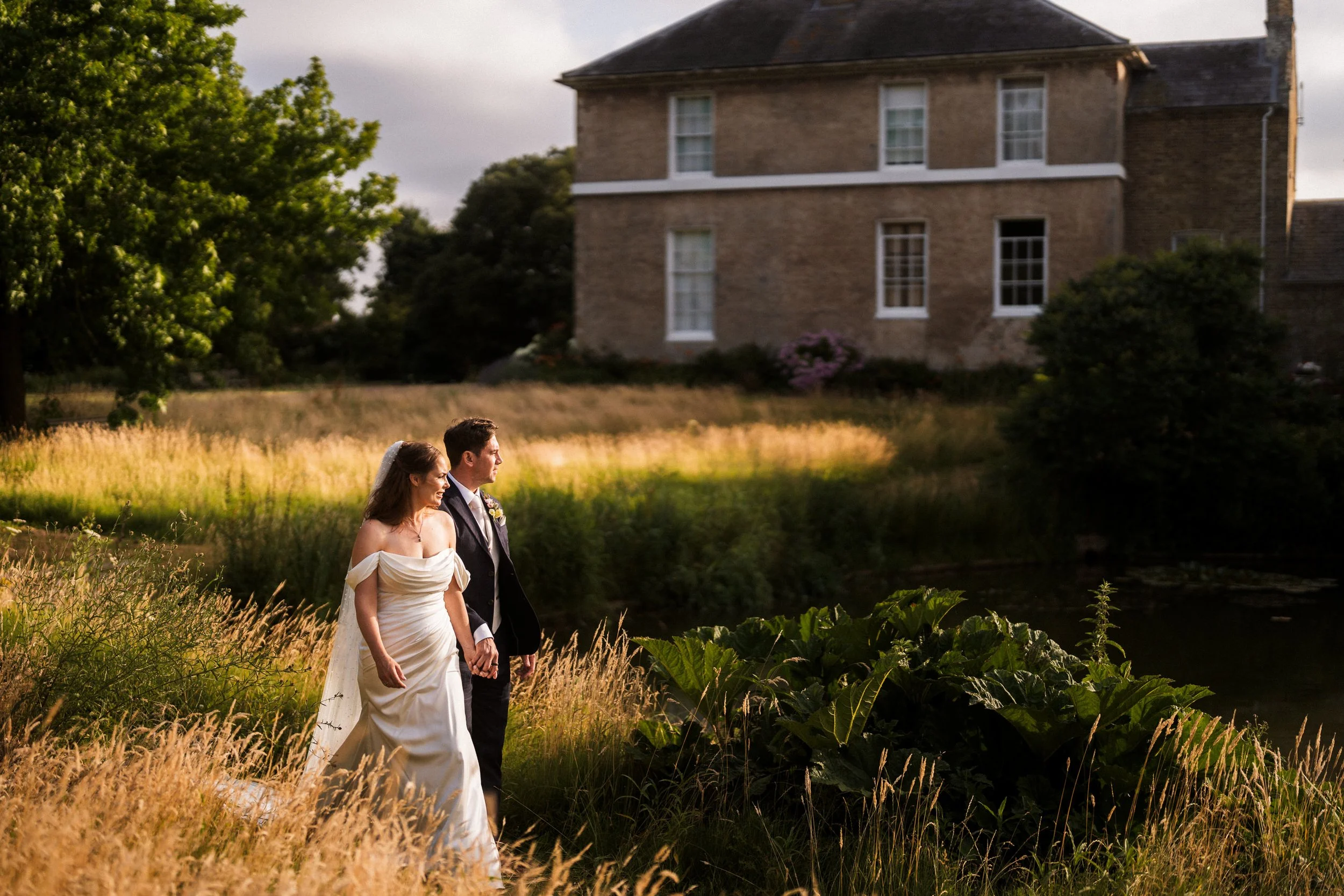 Newlywed couple walking through a sunlit meadow beside a historic brick house