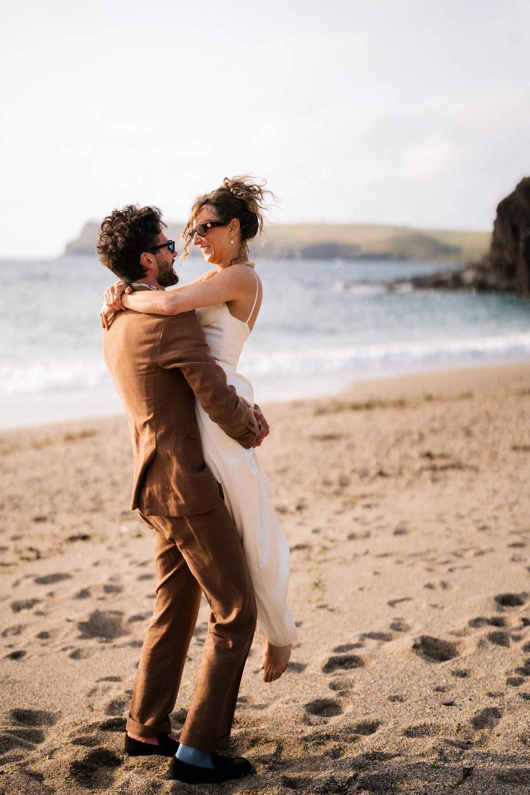 Smiling couple embracing on a sandy beach with ocean and rocky cliffs in the background