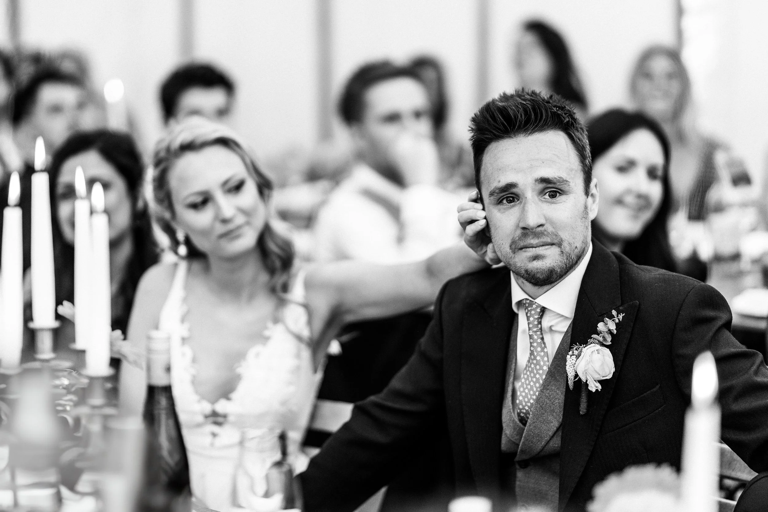 Black-and-white wedding reception photo of a groom in a suit and boutonniere seated among guests and candlelit tables.