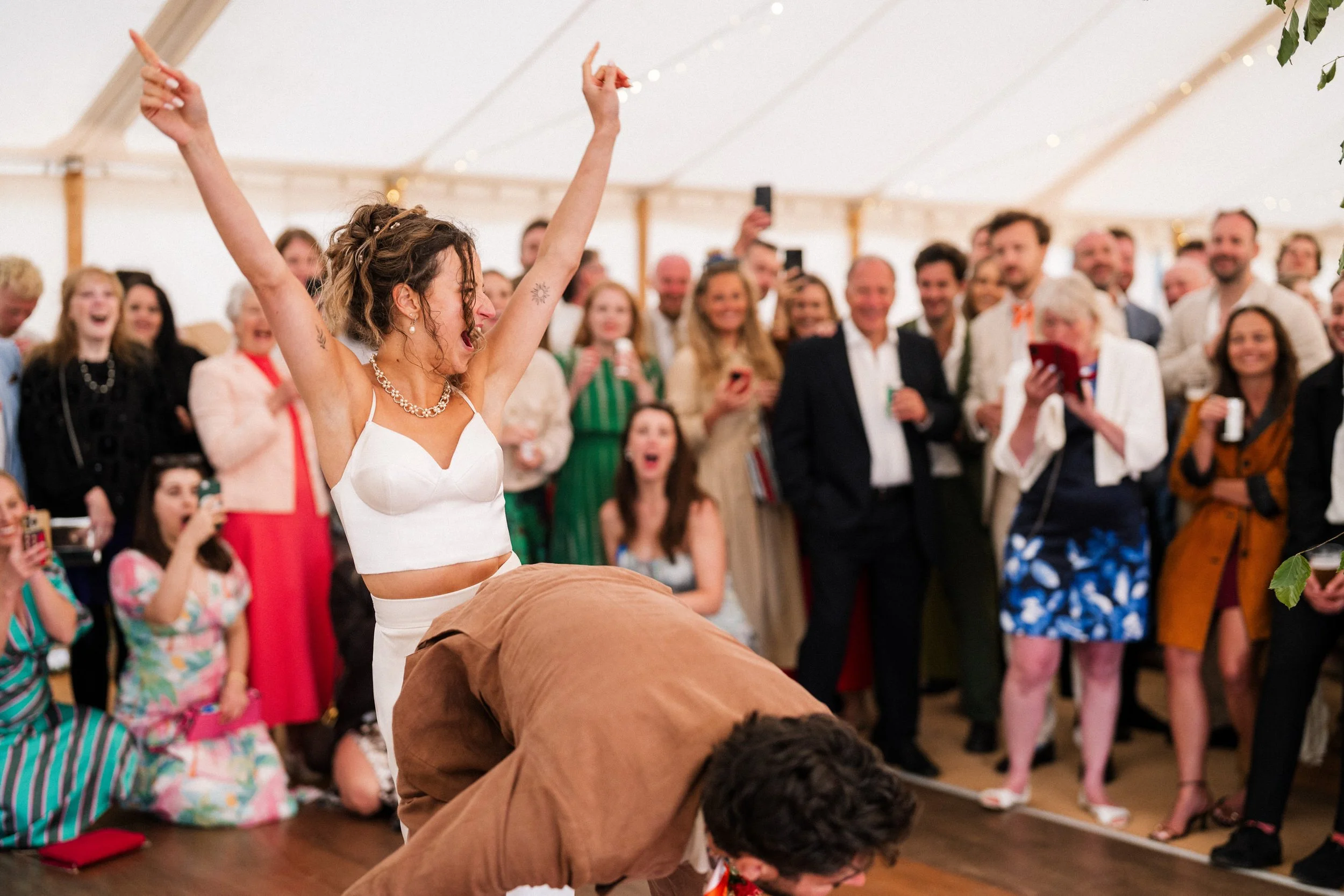 Bride dancing with raised arms as cheering wedding guests watch and take photos