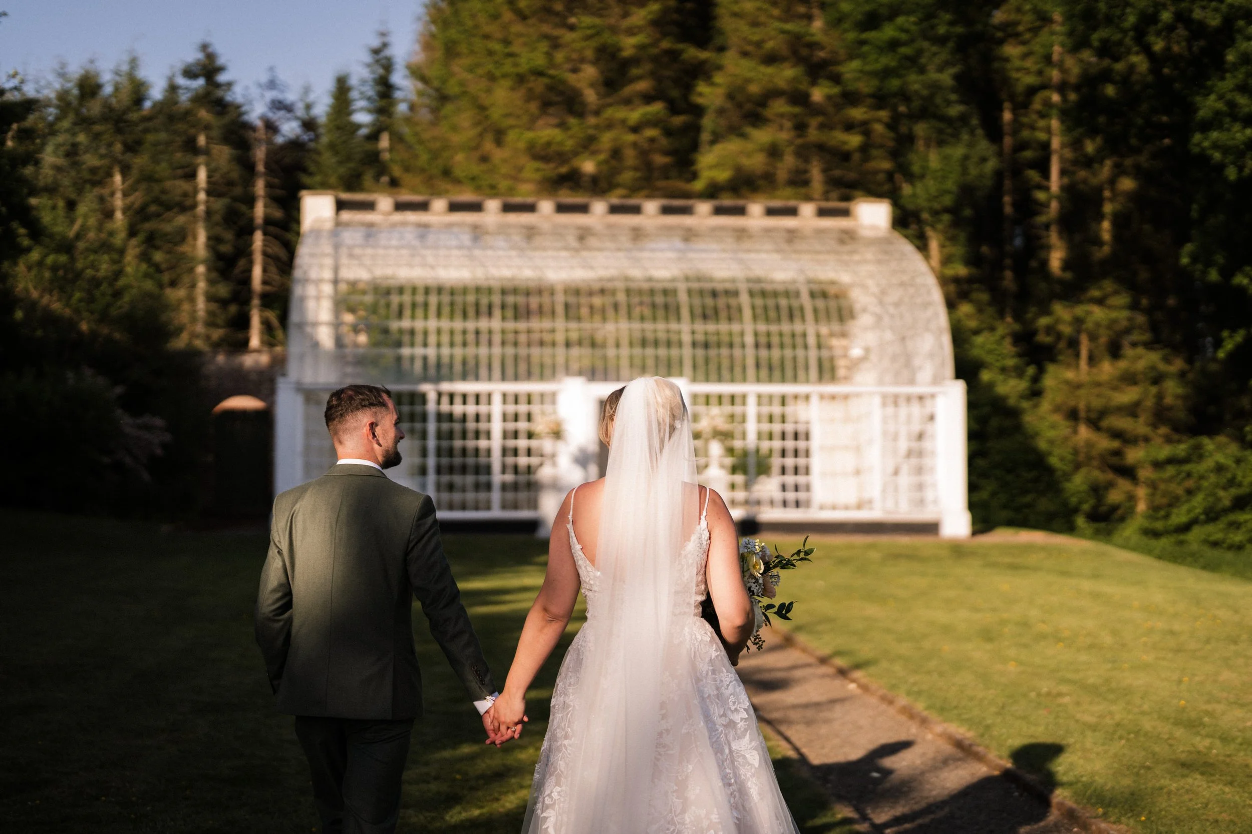 Bride and groom holding hands and walking toward a white garden gazebo surrounded by trees.