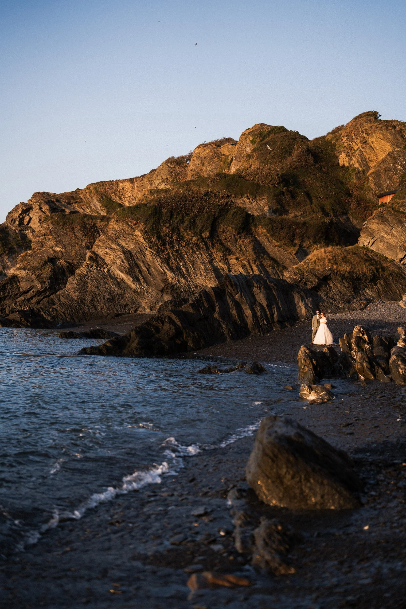 Bride and groom standing on a rocky black-sand beach beneath sunlit coastal cliffs
