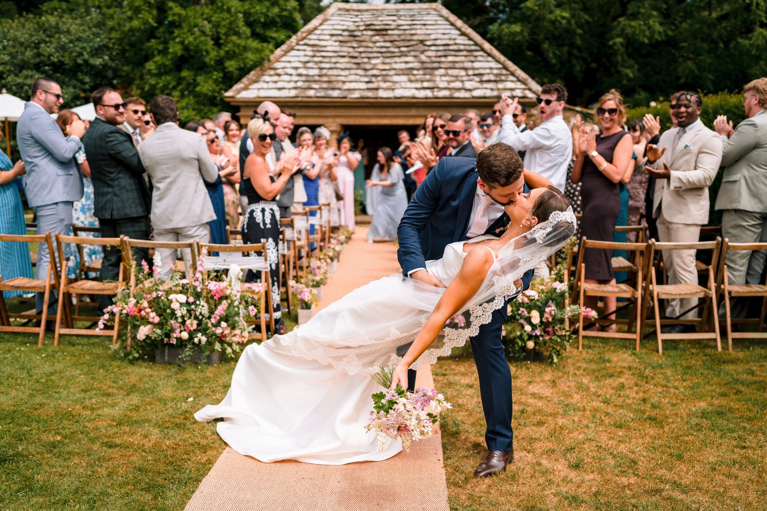 Newlywed couple kisses during an outdoor wedding ceremony as guests applaud around them.