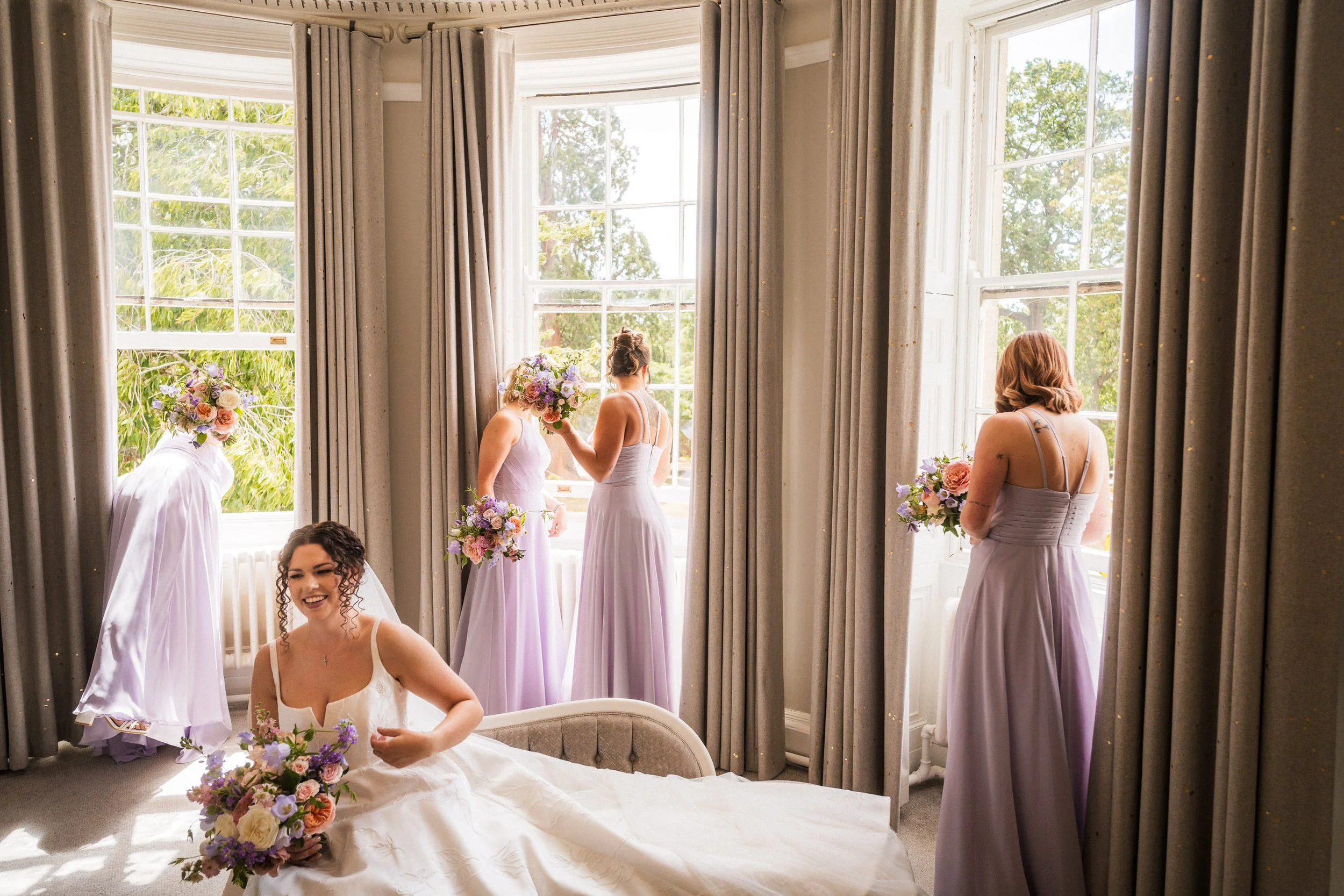 Bride and bridesmaids in lavender dresses holding bouquets in a bright bedroom with large windows