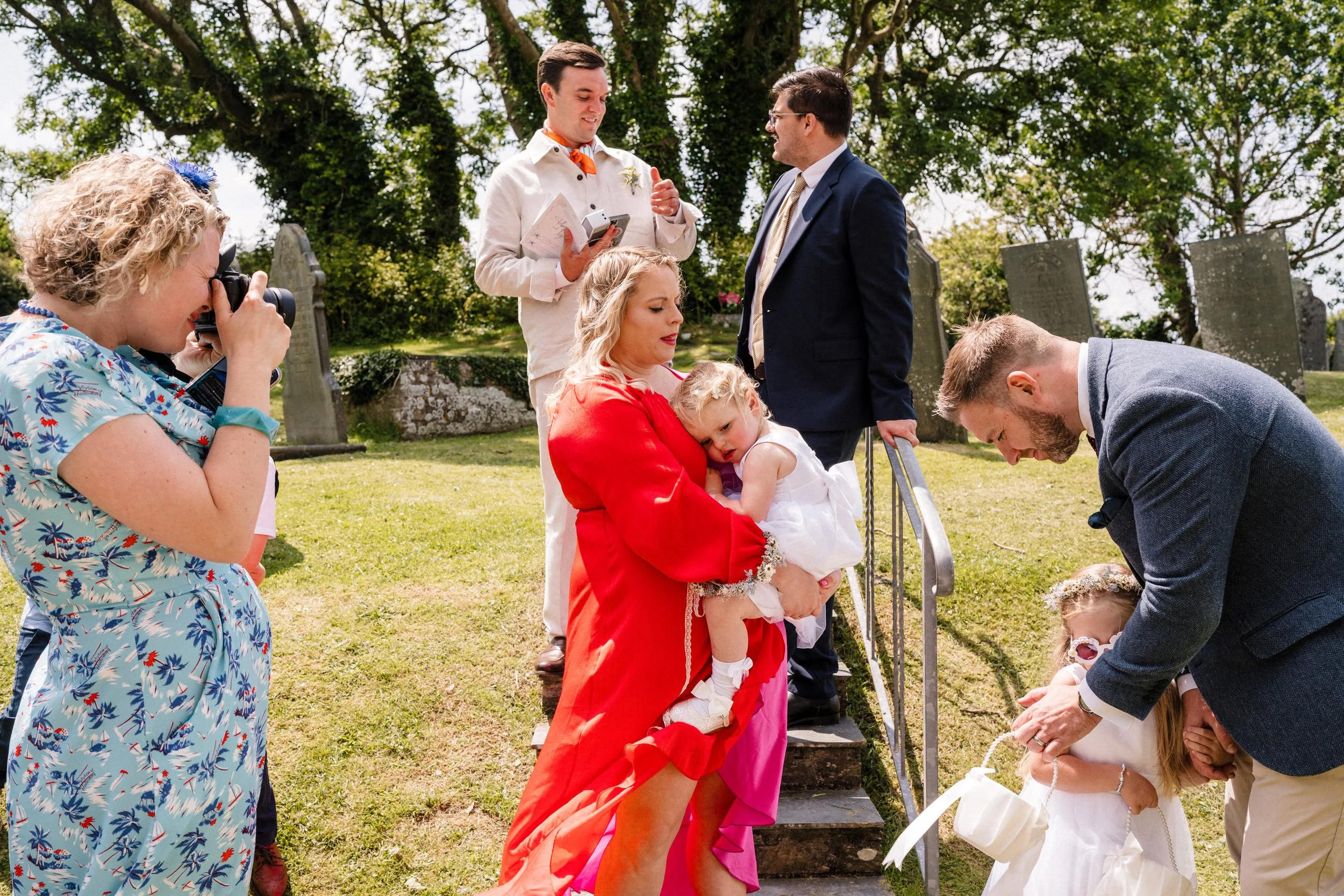 Wedding guests gather in a cemetery, with a photographer documenting a family carrying a toddler near gravestones.