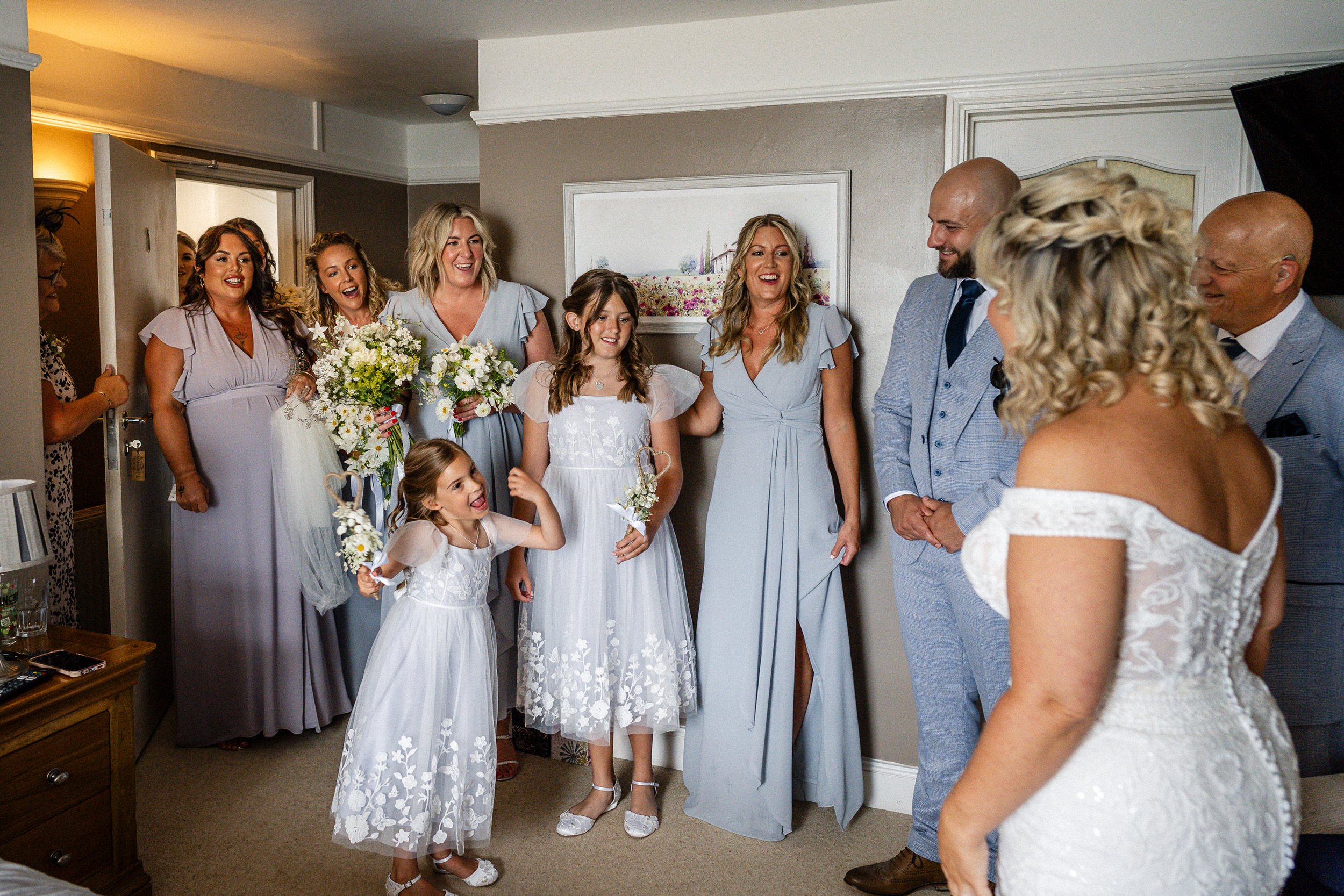 Wedding party gathered indoors with the bride, bridesmaids in lavender dresses, flower girls, and guests smiling around bouquets.