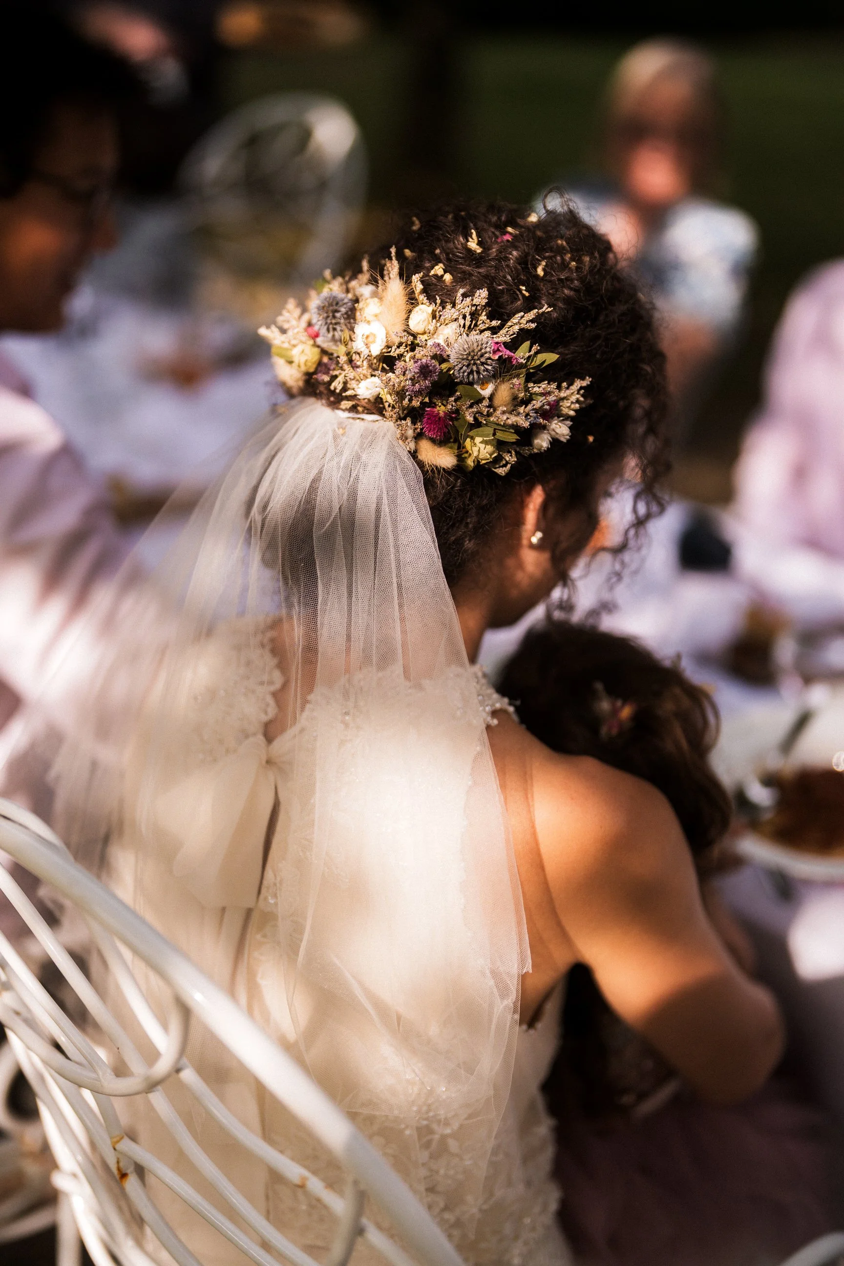 Bride with a dried floral hairpiece and long veil seated among wedding guests.