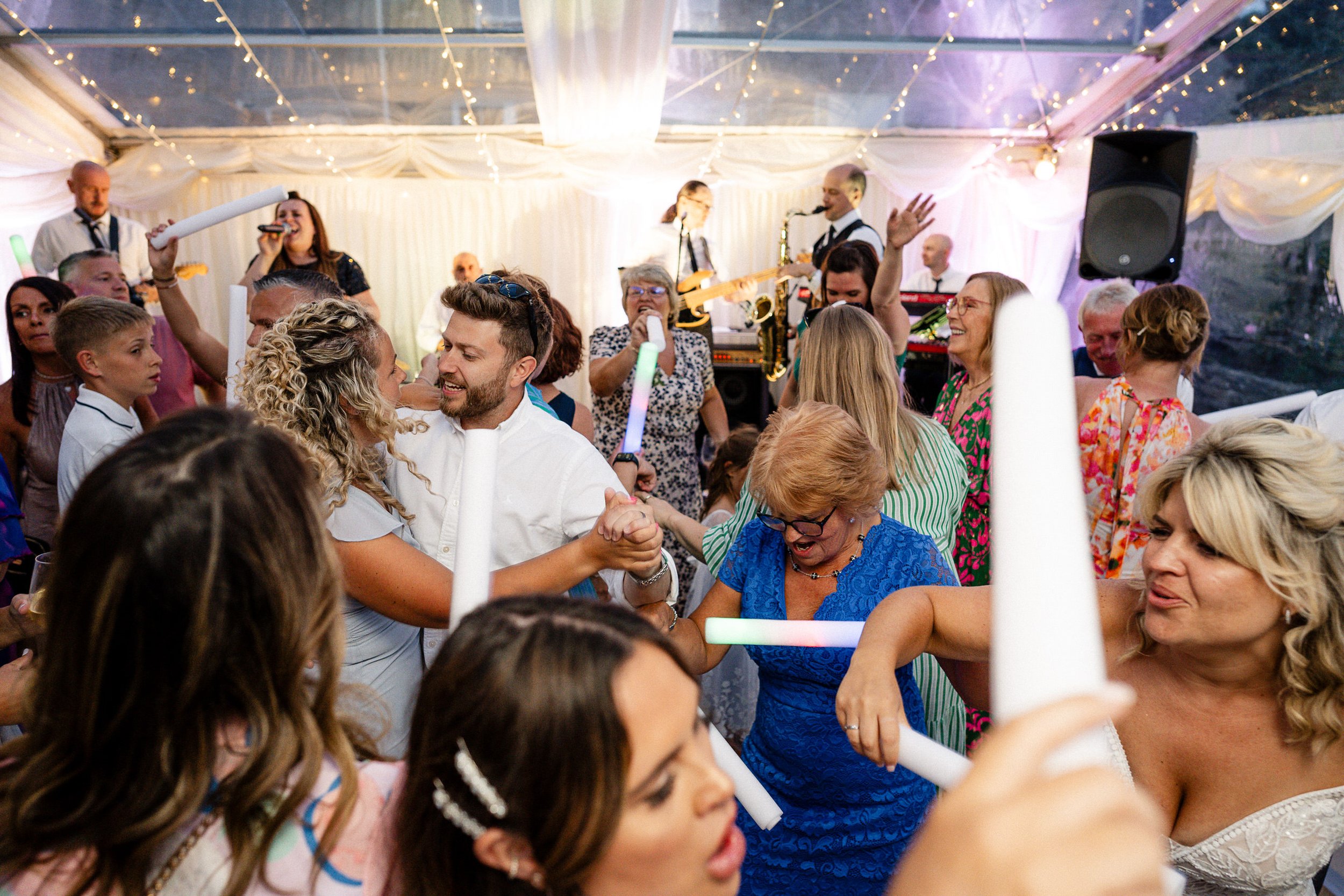 Wedding guests dancing with glow sticks as a live band performs under string lights at a reception.
