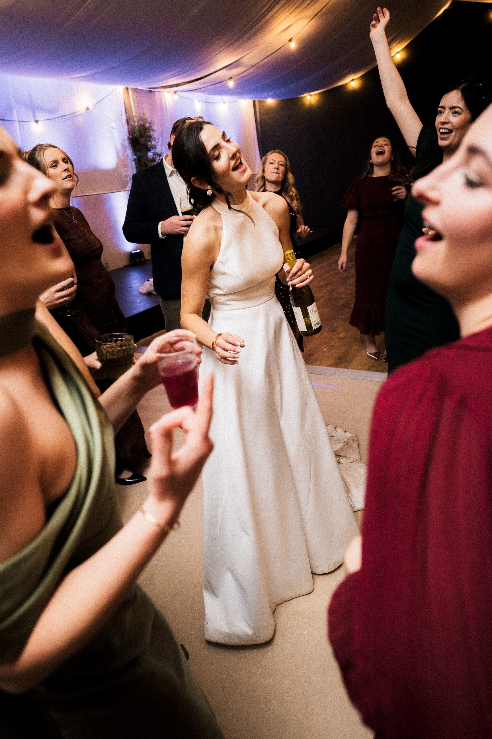 Bride holding a champagne bottle while dancing with guests at a warmly lit wedding reception