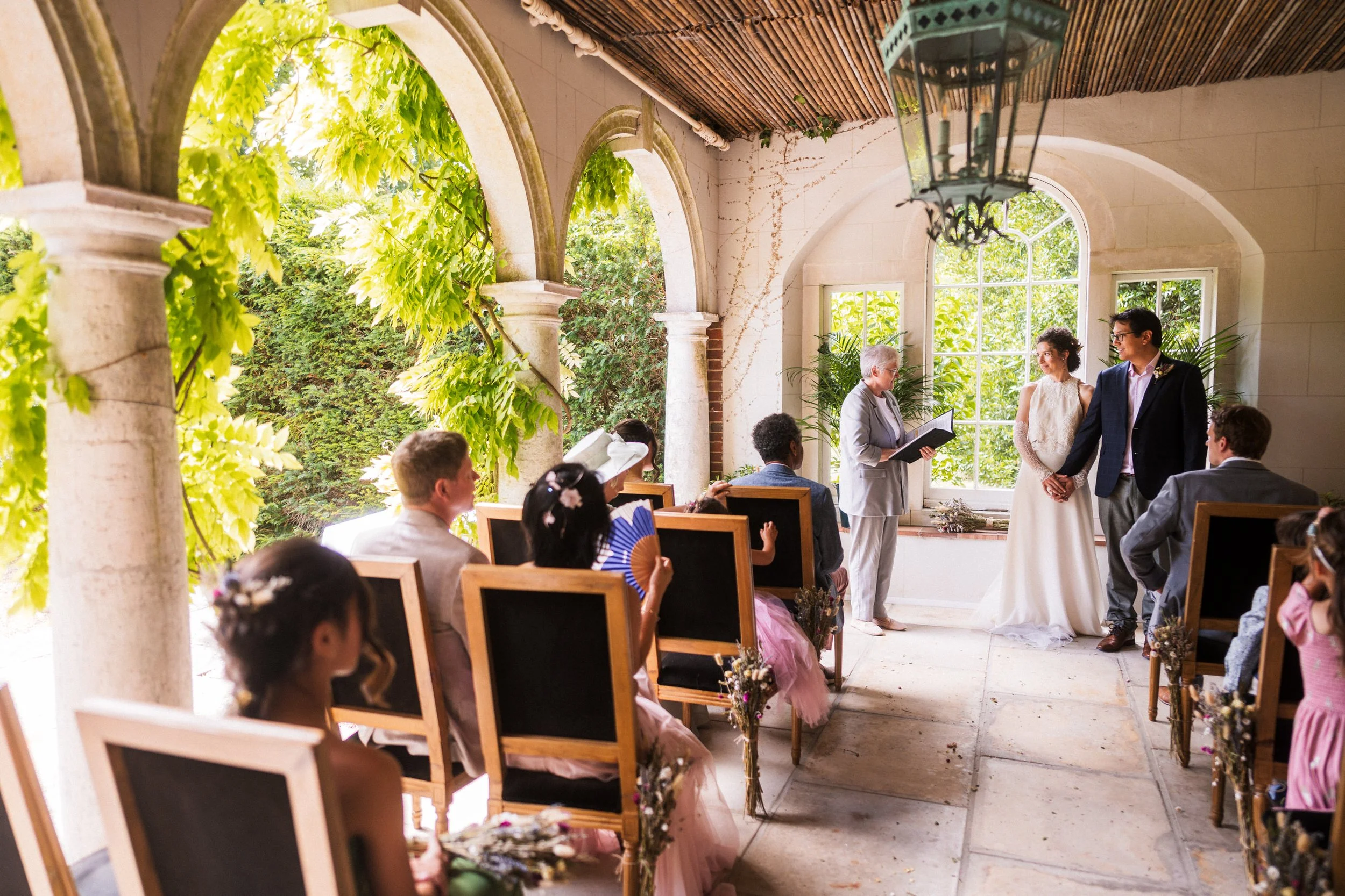 Wedding ceremony in a sunlit garden pavilion with guests seated as a bride and groom stand before an officiant.