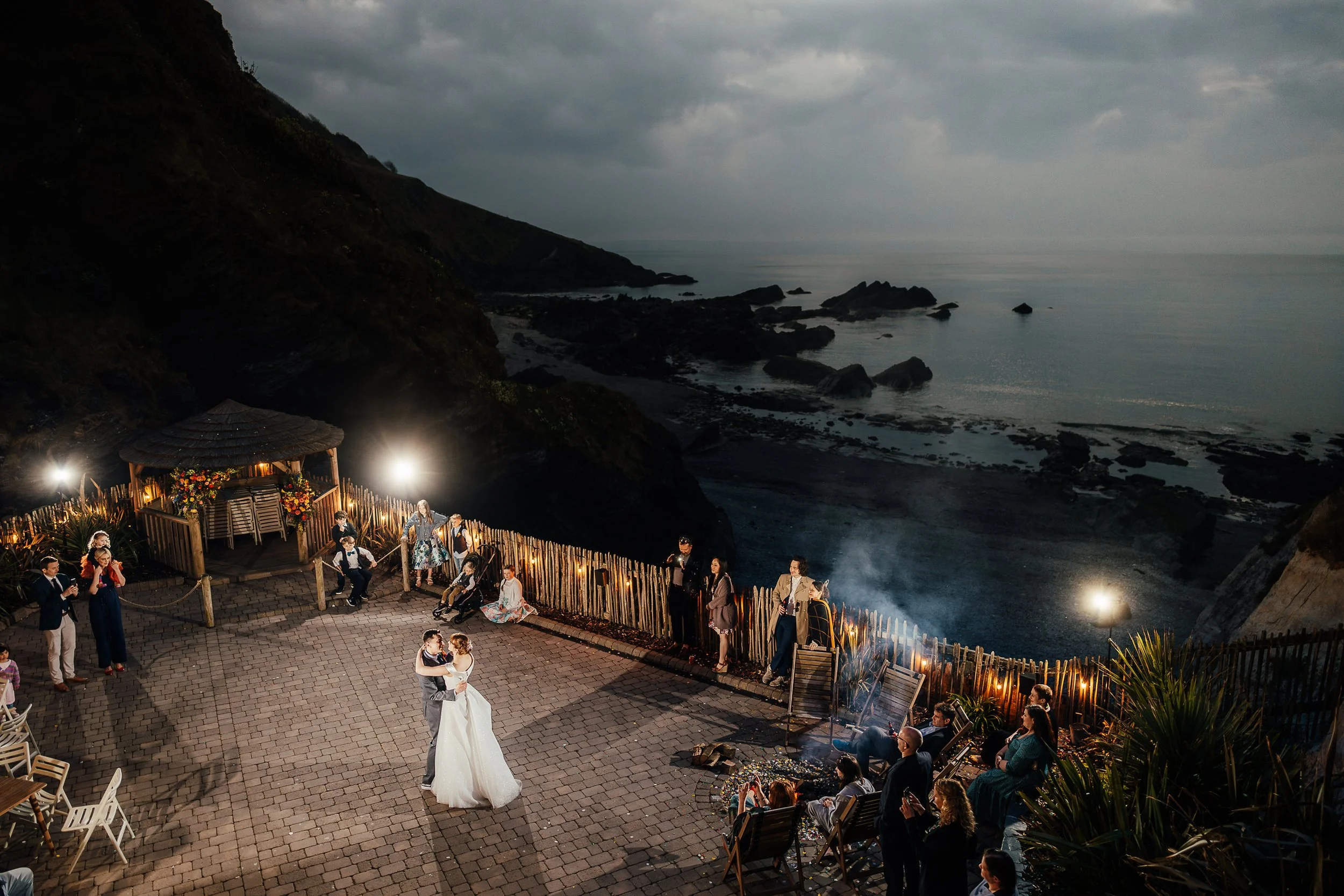 Outdoor wedding reception on a rocky ocean cliff at dusk, with the bride and groom dancing, guests seated around a fire pit, and a flower-decorated gazebo.