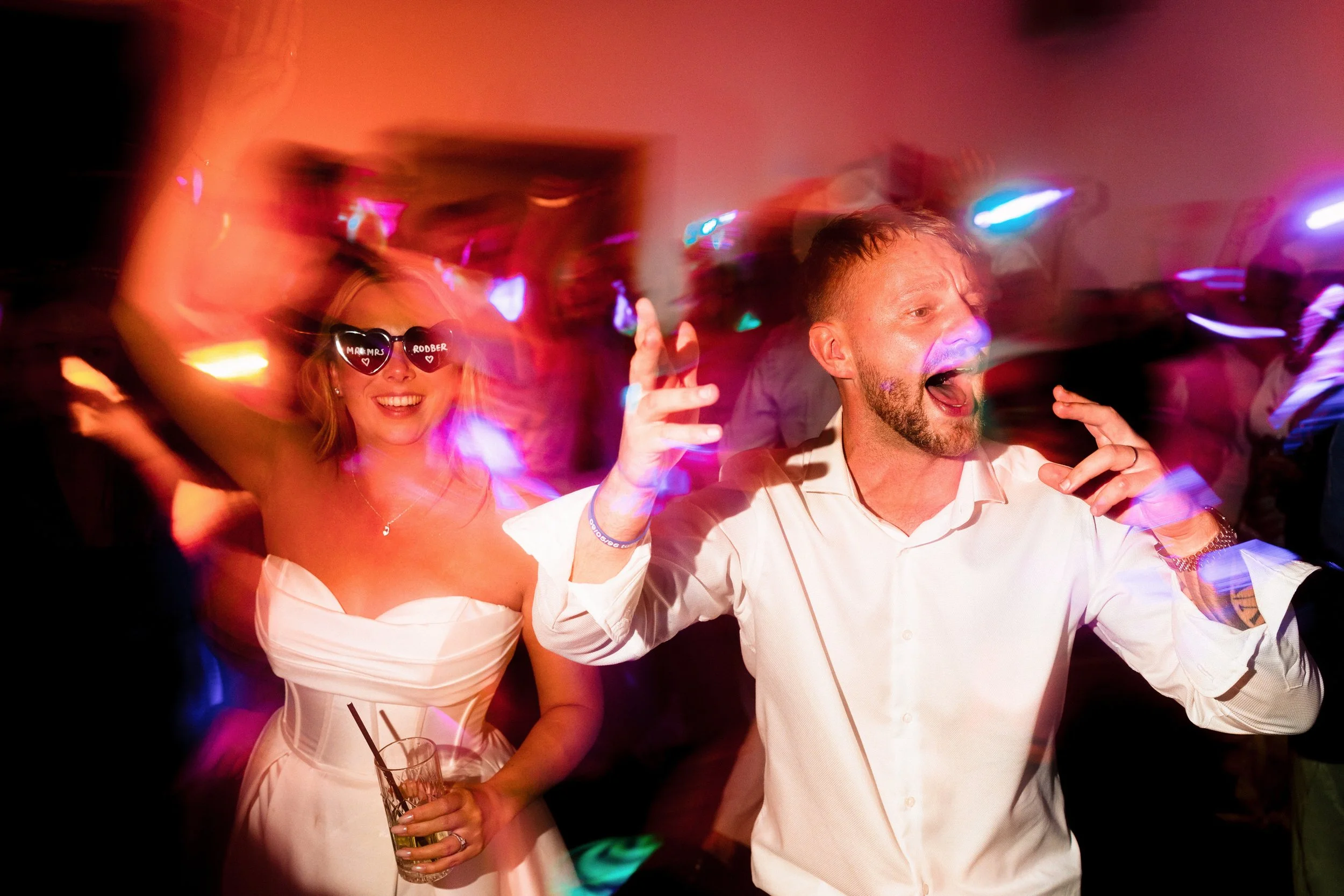 Couple dancing at a lively nightclub party under colorful neon lights, with the woman wearing heart-shaped sunglasses and holding a drink.