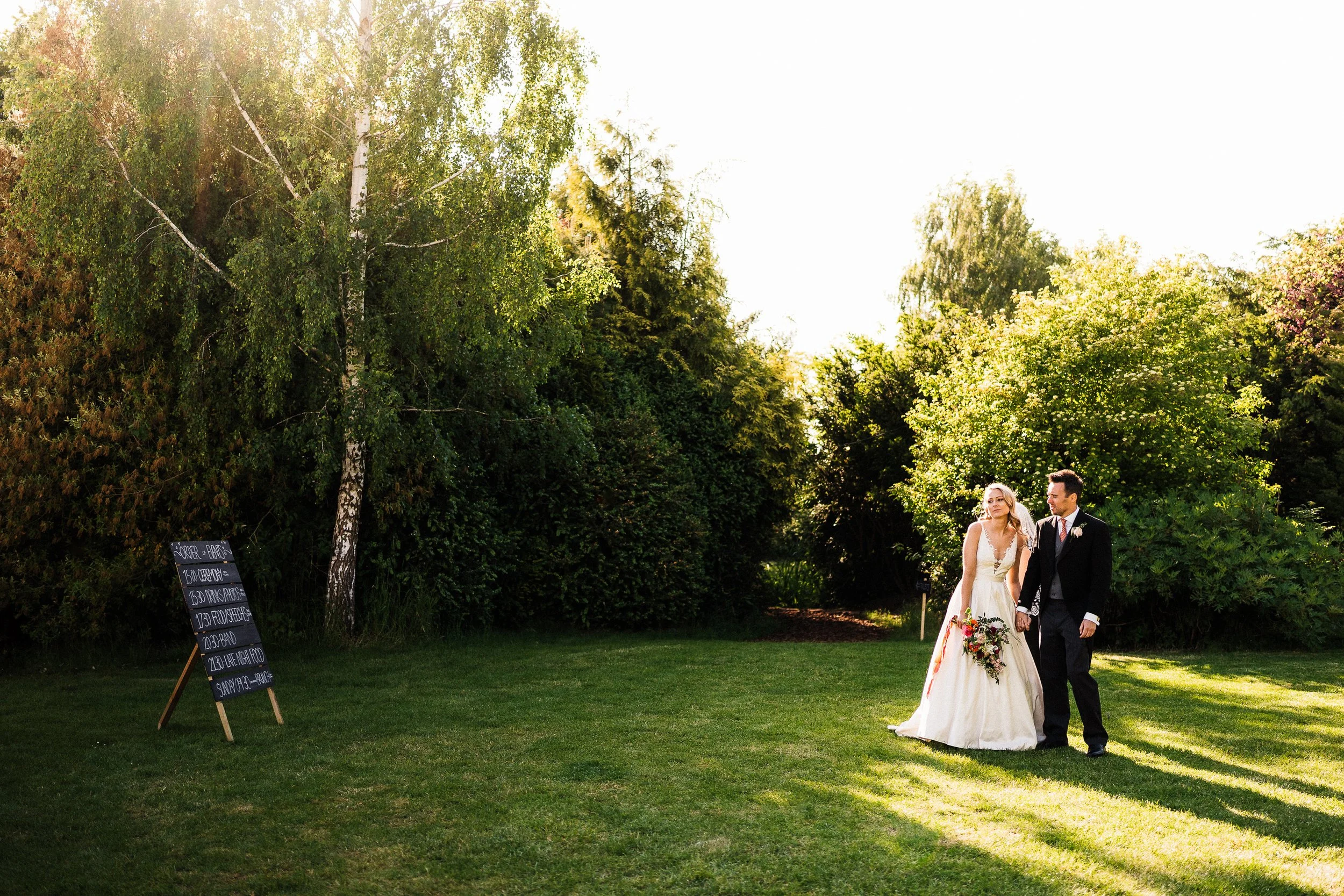 Bride and groom walking through a sunlit garden beside a wedding reception sign.