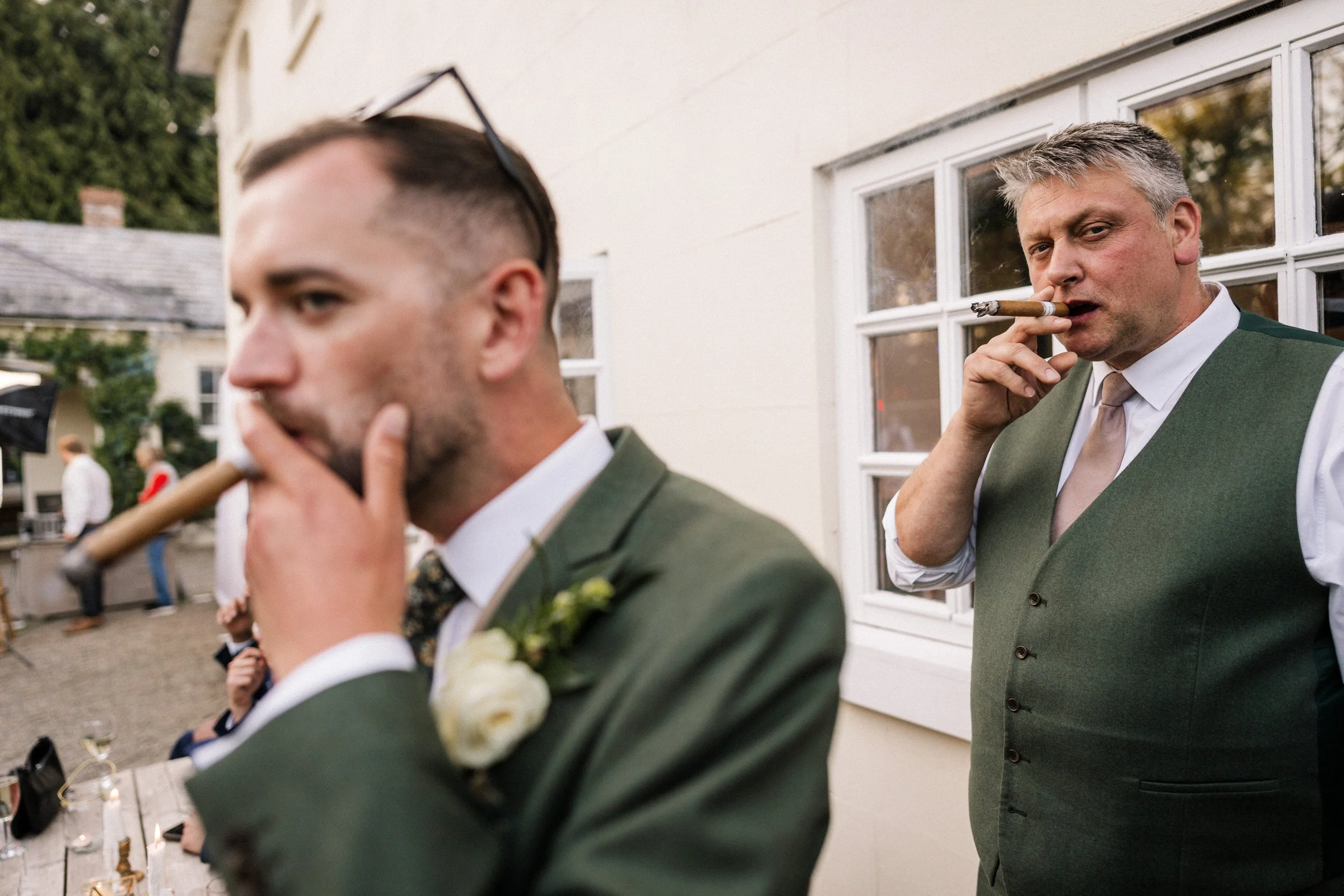 Two men in formal green suits smoking cigars at an outdoor wedding reception.