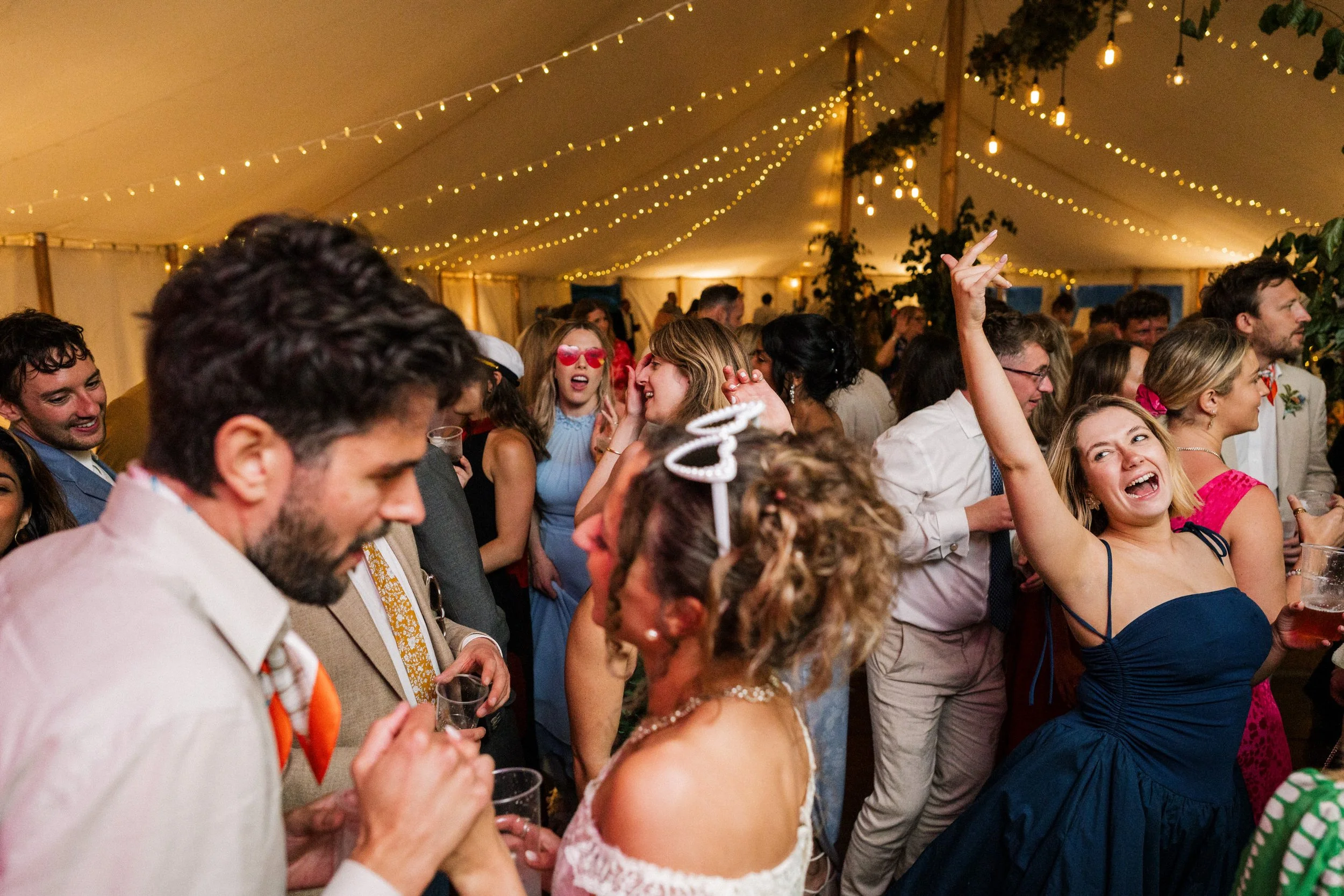 Crowded wedding reception with guests dancing and celebrating under string lights in a tent